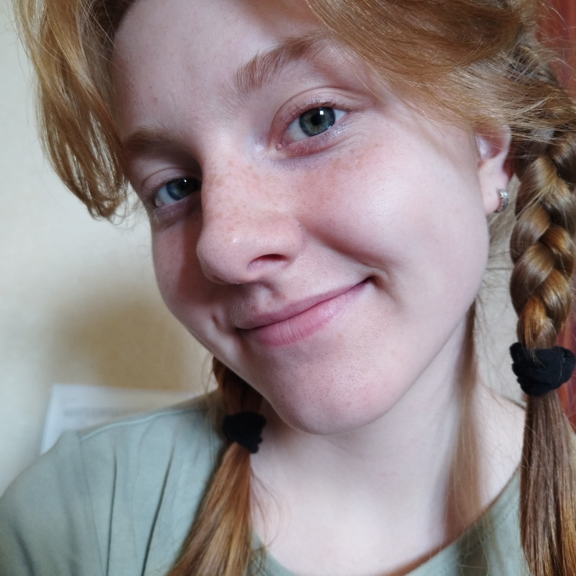 A close-up, slightly angled portrait features a young individual, likely female, looking directly at the camera with a gentle, closed-mouth smile. Her face, primarily the upper body from the chest up, fills most of the frame. She has fair skin, generously dusted with freckles across her nose and cheeks. Her eyes are a striking blue-green, reflecting the light.
Her hair is a reddish-blonde hue, styled into two braided pigtails, each secured with a black hair tie, visible on either side of her face. Loose strands frame her forehead and temples. Visible pores are present on her nose and chin area. She wears a light green or olive-toned shirt, part of which is visible on her shoulders and chest. A small, silver-toned hoop earring is worn in her right ear (viewer's left).
The background is softly blurred and appears to be a light-colored, possibly beige or off-white, indoor wall, suggesting an interior setting. The lighting is even and bright. The overall expression conveyed is one of contentment or pleasantness. There is no visible text in the image.