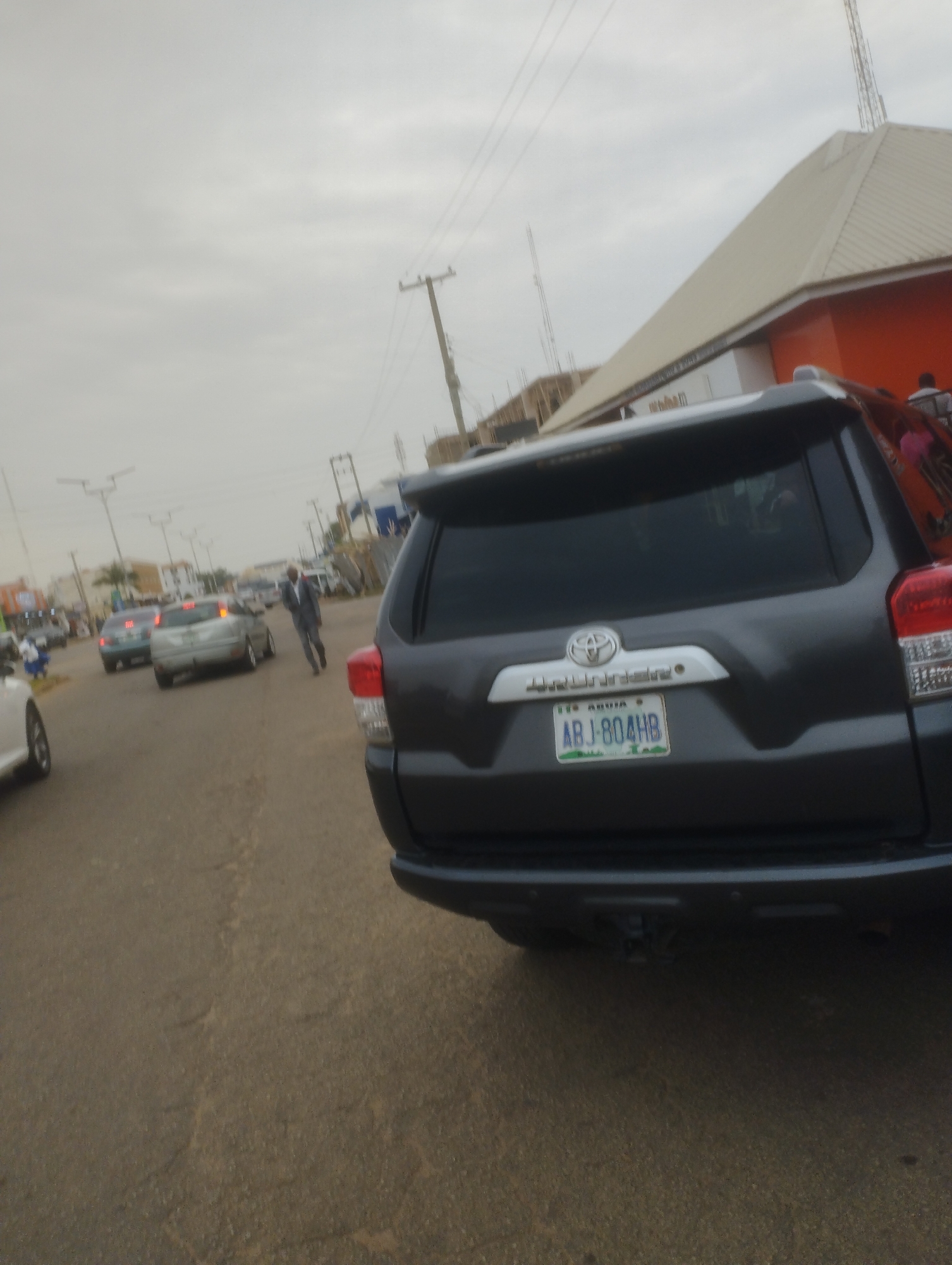 The image depicts a street scene in Jos, Nigeria, under an overcast sky. In the foreground, the rear of a dark gray SUV, possibly a Toyota 4Runner based on the emblem, is prominently featured. The SUV has a license plate that reads "ABJ-804HB" and appears to have a tow hitch at the rear.
The street is paved with asphalt and shows some signs of wear. Several other vehicles are visible on the road, including a silver car in the middle ground and a lighter colored vehicle on the far left. A man in a gray suit is walking on the road between the silver car and the SUV in the foreground. In the background, there are buildings, including one with a distinctive peaked roof that is light-colored with an orange section. Utility poles with overhead wires are also visible. The overall impression is of a typical urban environment on a cloudy day.