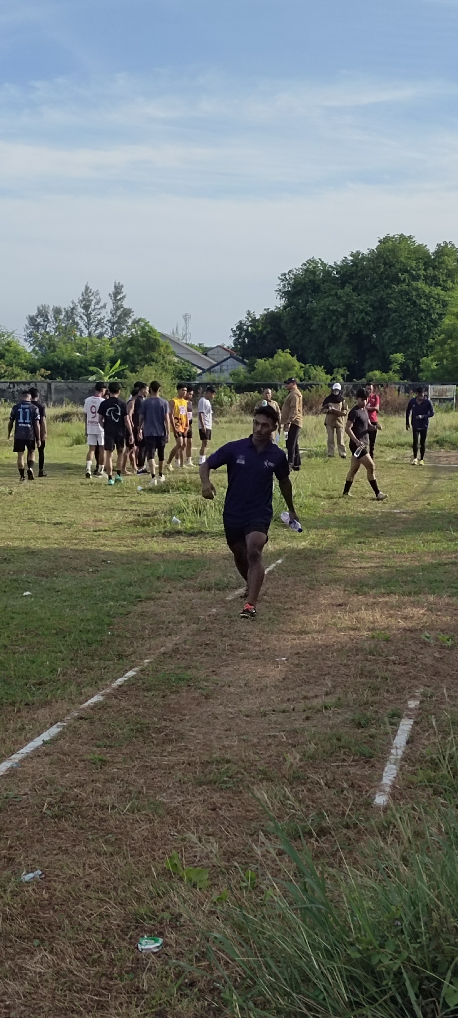 The image depicts a group of young men on a grassy field, likely a sports ground, under a partly cloudy sky. In the foreground, a young man in a dark blue polo shirt and dark shorts is running towards the viewer, holding a clear plastic water bottle in his right hand. He is wearing athletic shoes. Further back, a group of men are gathered, some wearing athletic attire, including numbered jerseys. One man in the background wears a tan jacket and a cap, possibly an instructor or observer. The environment is a rural or semi-rural setting with trees and a low-rise building visible in the distance. The ground is covered in dry grass with some green patches and is marked with white lines, suggesting a track or court. The presence of litter, such as discarded paper cups, is noticeable on the ground. While the exact location is not explicitly identified, the context suggests it could be an outdoor sports facility in Banda Aceh, Indonesia, given the typical weather and vegetation.