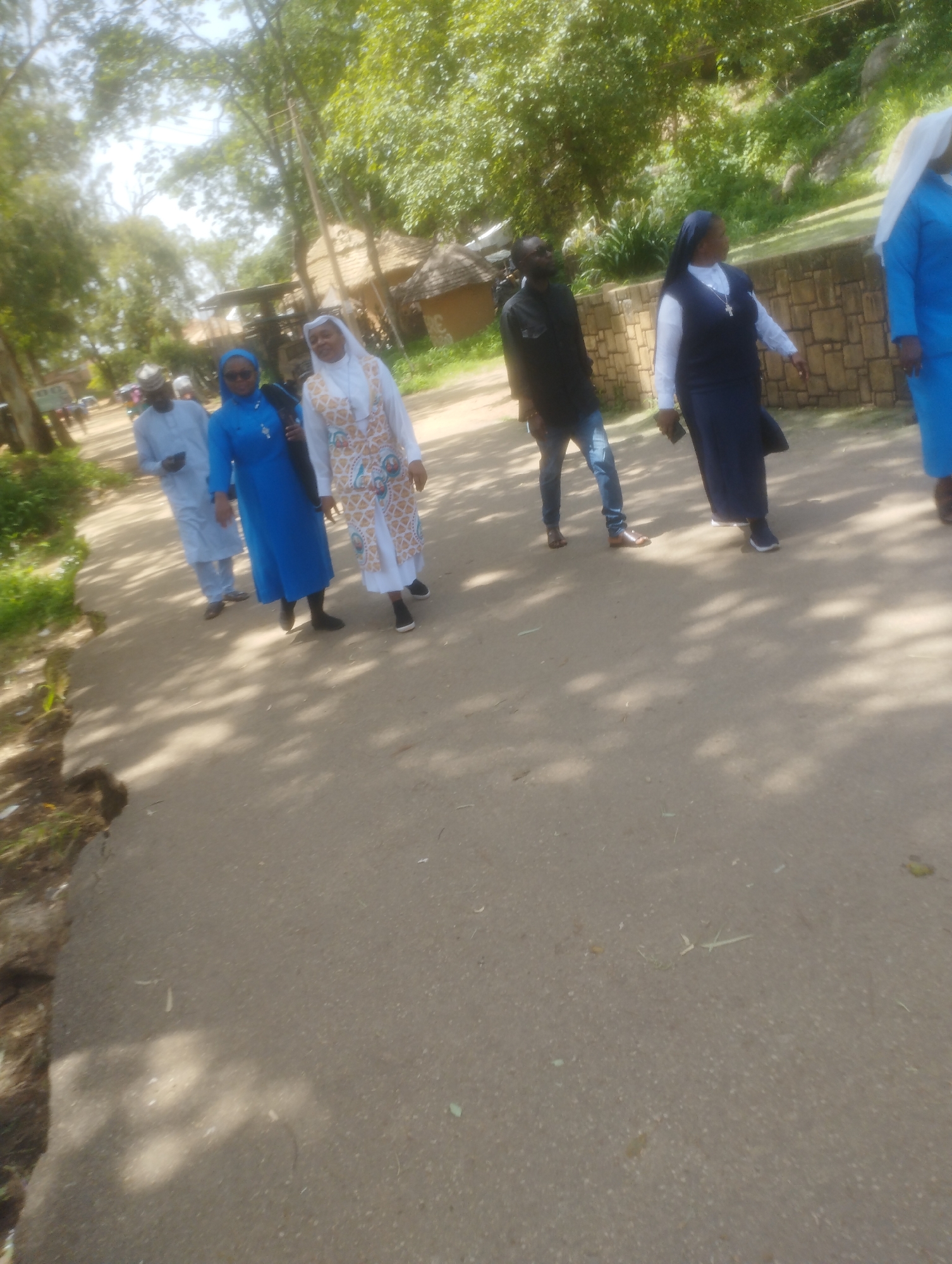 A group of individuals are walking on a paved road in Jos, Nigeria. The group includes three women in religious attire, two of whom are wearing blue habits with white veils and one in a white and patterned dress with a white headscarf. Two men are also present, one in a black shirt and jeans, and another in a light blue traditional outfit. The environment is outdoors with trees and vegetation visible in the background, suggesting a natural or park-like setting. Sunlight filters through the trees, casting shadows on the road. There are also some structures visible, possibly buildings or shelters, with thatched roofs. The overall scene depicts a casual outdoor gathering or procession.