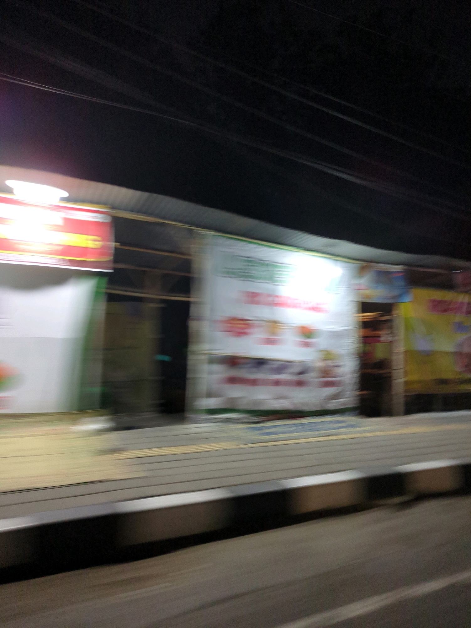 This is a blurry, horizontal, nighttime, street-level photograph taken from a moving vehicle in Colomadu, Indonesia. The image captures a row of illuminated storefronts and advertisements lining a sidewalk, with a dark sky and overhead wires visible at the top. The motion blur suggests a fast shutter speed, creating streaks of light and indistinct forms. On the left, a red and yellow sign is partially visible. To its right, a large white banner with green lettering and images of food items is prominently displayed. Further to the right, another banner, this one yellow with red lettering and images of food or drinks, is also visible. The buildings appear to be single-story structures with corrugated metal roofs. The sidewalk is paved and has a raised edge. The overall impression is of a bustling commercial area at night, seen in transit.