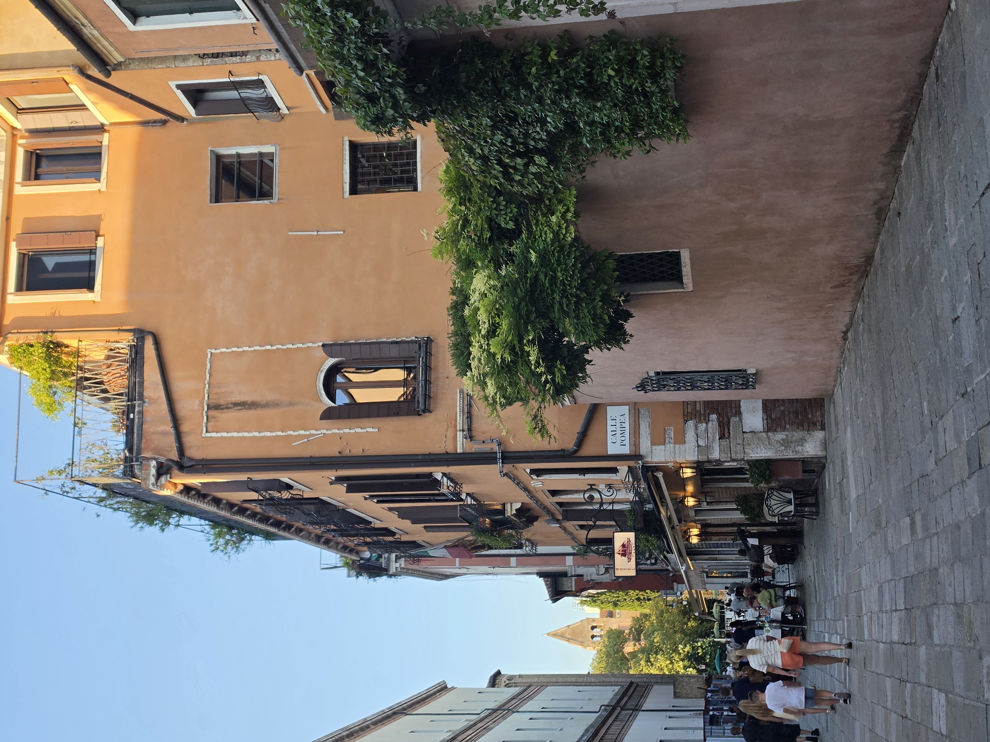 The image shows a narrow street in Venice, Italy, lined with tall, narrow buildings. The buildings are predominantly orange and pink, with shutters on the windows. There are some plants growing on the buildings, and a rooftop garden is visible on one of the buildings. The street is paved with cobblestones, and there are a few people walking in the street. On the right side of the street, there is a sign that reads "CALLE POMPEA." There are also some outdoor seating areas for restaurants. The overall impression is of a charming, old-world street scene.