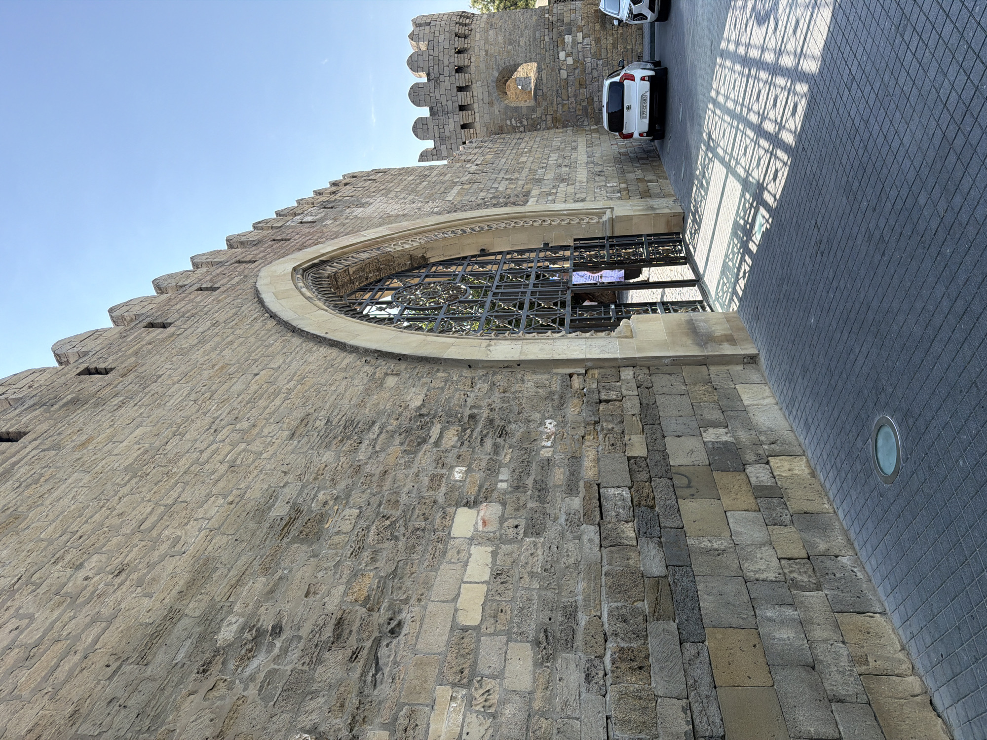 A wide-angle outdoor shot captures a portion of the stone walls of the Maiden Tower in Sabail Raion, Azerbaijan, on a sunny day. The wall on the left is constructed of rough-hewn, tan and gray stones of various sizes, with a visible arched gateway featuring ornate black metalwork. Beyond the gate, figures are partially visible. To the right, the wall continues, transitioning to a cylindrical tower with crenellations and an arched window. Two white cars are parked in front of the wall; the one on the left displays a license plate with the characters "77 CC 683." The ground is paved with dark gray square tiles arranged in a diamond pattern, and intricate shadows from the gateway's metalwork are cast across it. The sky above is a clear, bright blue.