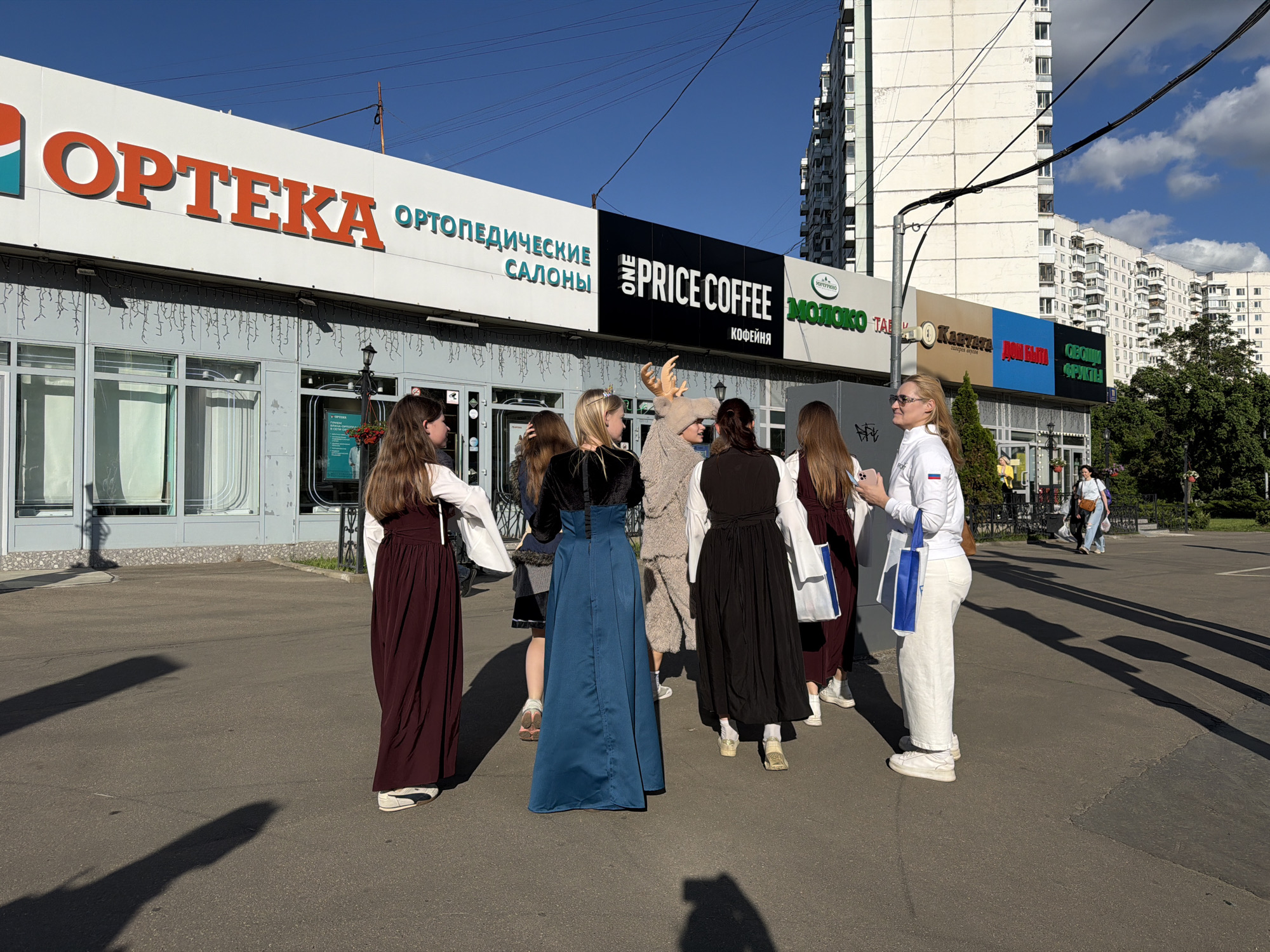 The image shows a group of people standing in front of a commercial building in Khimki, Russia. The building has several storefronts with signs in Russian, including "OPTEKA" (pharmacy), "Ortopedicheskiye Salony" (Orthopedic Salons), "One Price Coffee," "MOLOKO" (Milk), and others.
There are several people standing in a group in front of the building. Most appear to be young women wearing long, simple dresses or skirts with white blouses. One person is wearing an animal costume with antlers. One person is wearing white pants and a white jacket. They appear to be having a conversation.
The location is urban, with apartment buildings visible in the background. The sky is blue with some clouds. The ground is paved. Shadows are cast by the building and the people, indicating a sunny day. There are other pedestrians in the background.