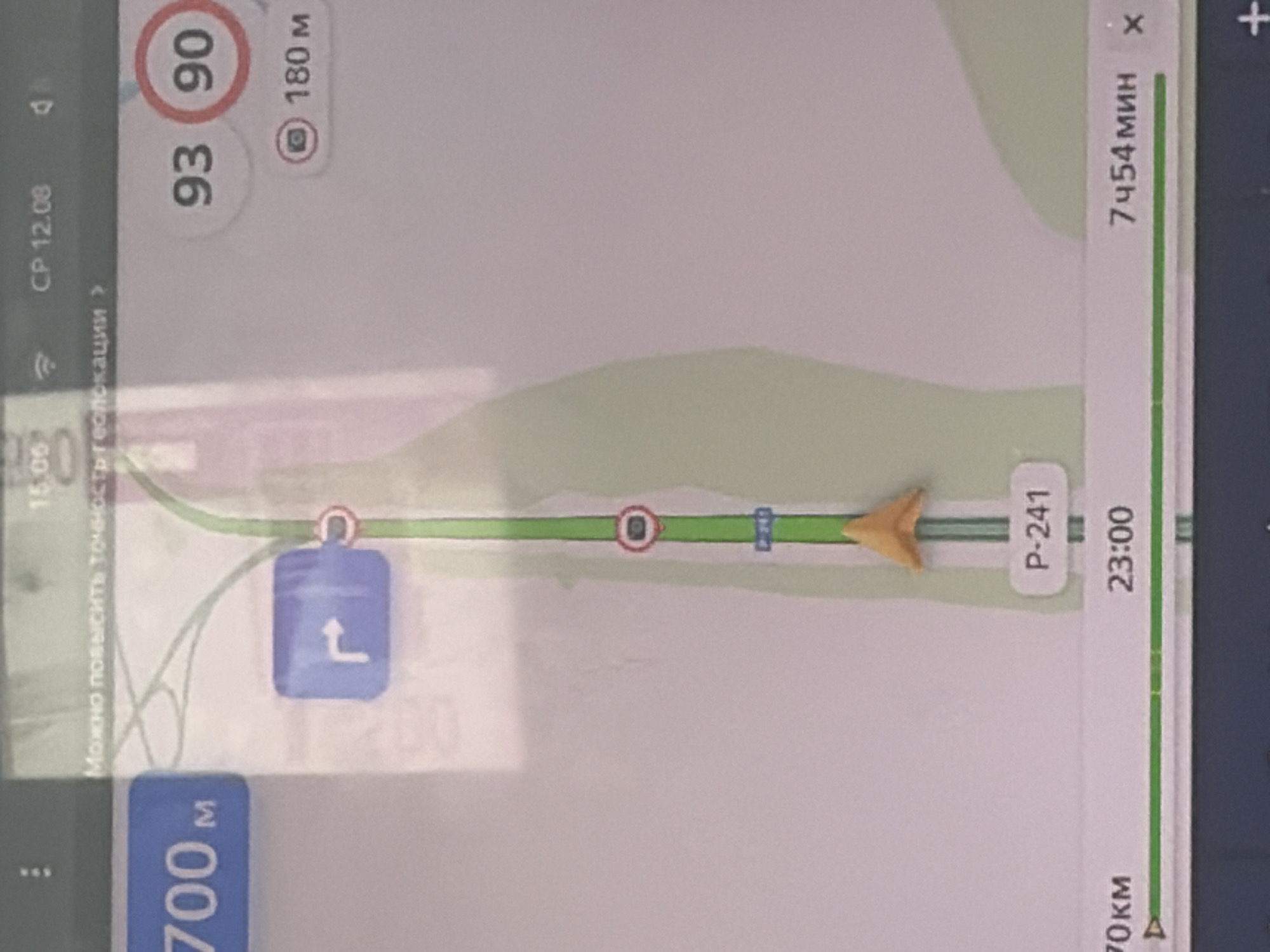 The image shows a screenshot from a car navigation system, likely displaying a map of Vershiny, Russia. On the screen, there is a route highlighted in green, indicating the path to be followed. Several icons and text overlays provide information about the navigation.

At the top of the screen, the time "CP 12.08" is visible, along with a signal strength indicator. Below this, there is a message in Russian: "Можно повысить точность геолокации" which translates to "Can improve geolocation accuracy."

To the left of the screen, "700m" is displayed, indicating the distance to a point or turn. A blue sign with a white arrow indicating a turn to the right is also visible. Further down the route, a circular sign with a red border and a number "90" inside indicates a speed limit of 90. Next to it, "93" is displayed, likely the current speed. A camera icon with "180m" suggests a speed camera located 180 meters ahead.

Along the bottom of the screen, "70км" (70 km) and "23:00" are displayed, possibly representing distance traveled or a destination. To the right, "7ч54мин" (7 hours 54 minutes) indicates the estimated time of arrival or remaining travel time. An "x" icon is present, which typically signifies closing or dismissing the information. A yellow directional arrow points upwards on the map, indicating the current direction of travel. The road number "P-241" is also visible.

The environment appears to be a road with some greenery surrounding it, suggesting a rural or suburban setting. The overall presentation is typical of a GPS navigation interface on a digital display.