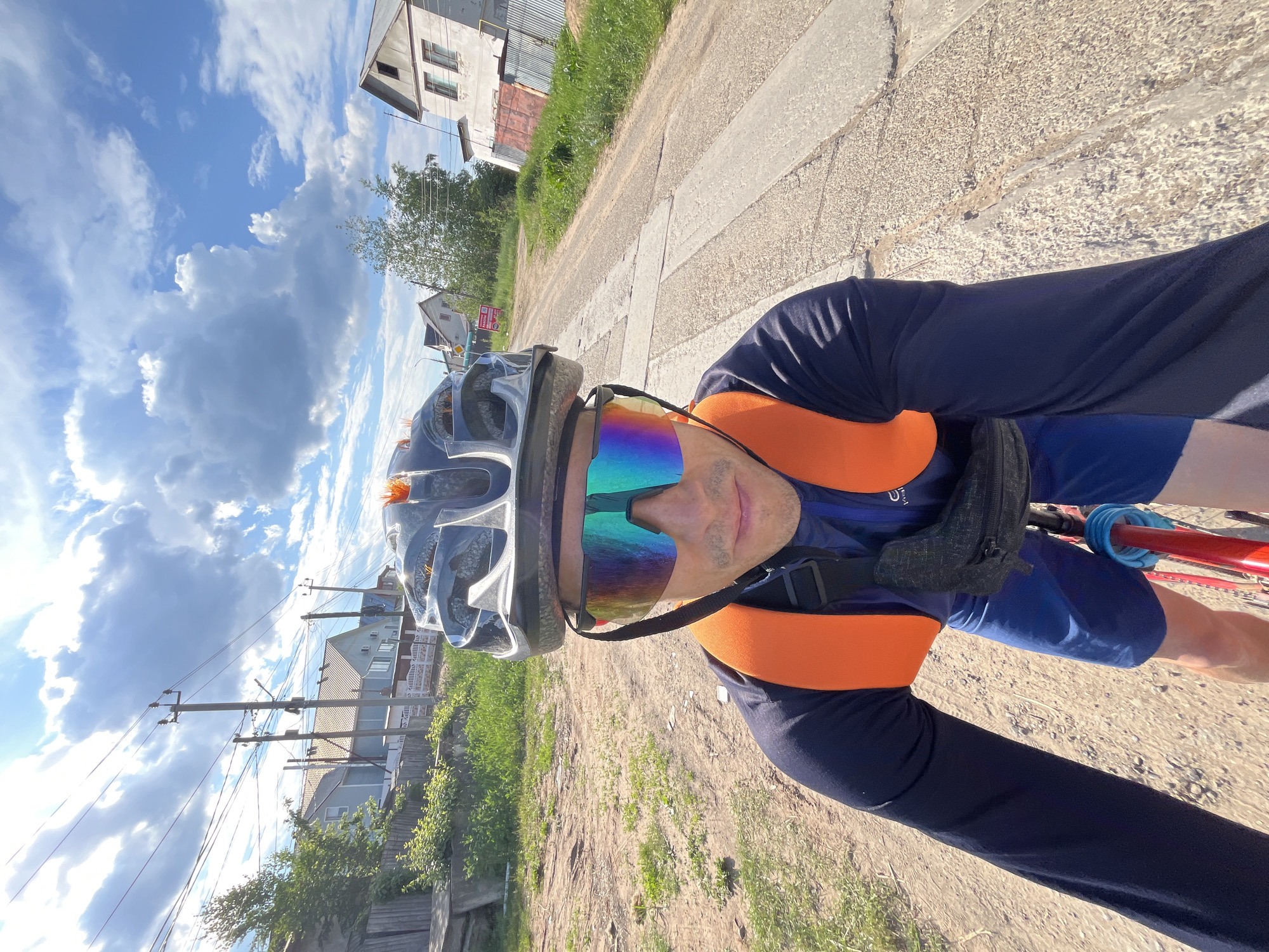 A man wearing a helmet and brightly colored cycling glasses is taking a selfie while riding a bicycle on a dirt road in the city of Chaikovsky, Russia. He is wearing a dark blue long-sleeved shirt, blue shorts, and an orange backpack. The road is lined with grass and houses. The sky is blue with white clouds.