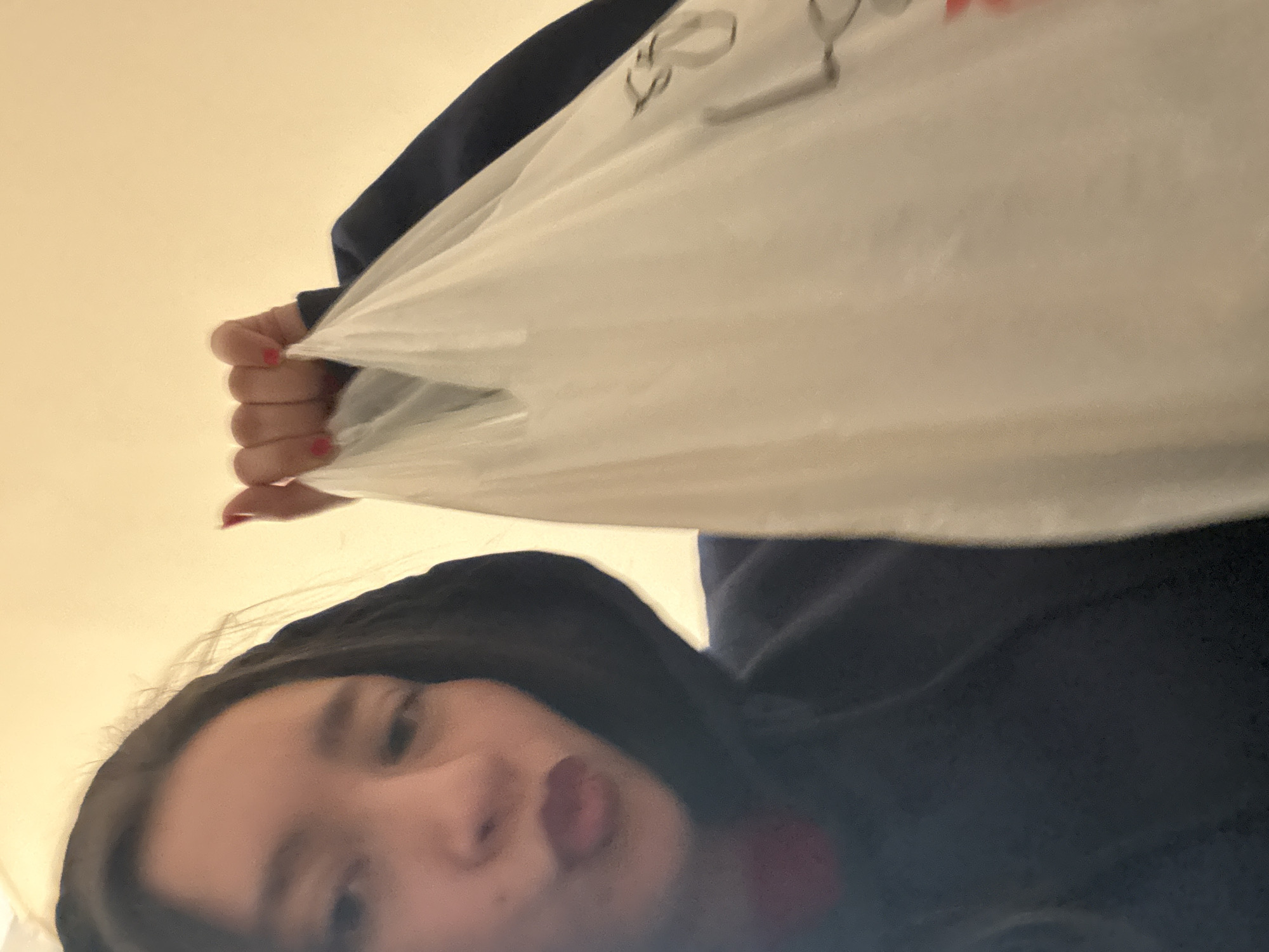 A low-angle shot shows a person holding a white plastic shopping bag overhead. The person has dark hair and is wearing a navy blue hooded sweatshirt. Red nail polish is visible on the fingers holding the bag. The face of the person is visible with an upward gaze. The background is a plain off-white wall. The city of Sofia, Bulgaria is not discernible from this image.