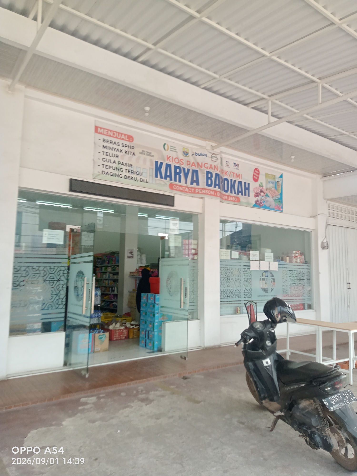 The image shows the exterior of a shop called "KARYA BAROKAH" in Jambi, Indonesia. A black motorcycle is parked in front of the shop. The shop appears to be a convenience store, with shelves of goods visible through the glass doors. A sign above the entrance lists items for sale, including rice, cooking oil, eggs, sugar, flour, and frozen meat. A person in a black hijab is partially visible inside the store. The shop's facade is white, and the roof is made of corrugated metal. A wooden table is set up outside the shop. The timestamp on the image indicates it was taken on September 1, 2026, at 2:39 PM.
