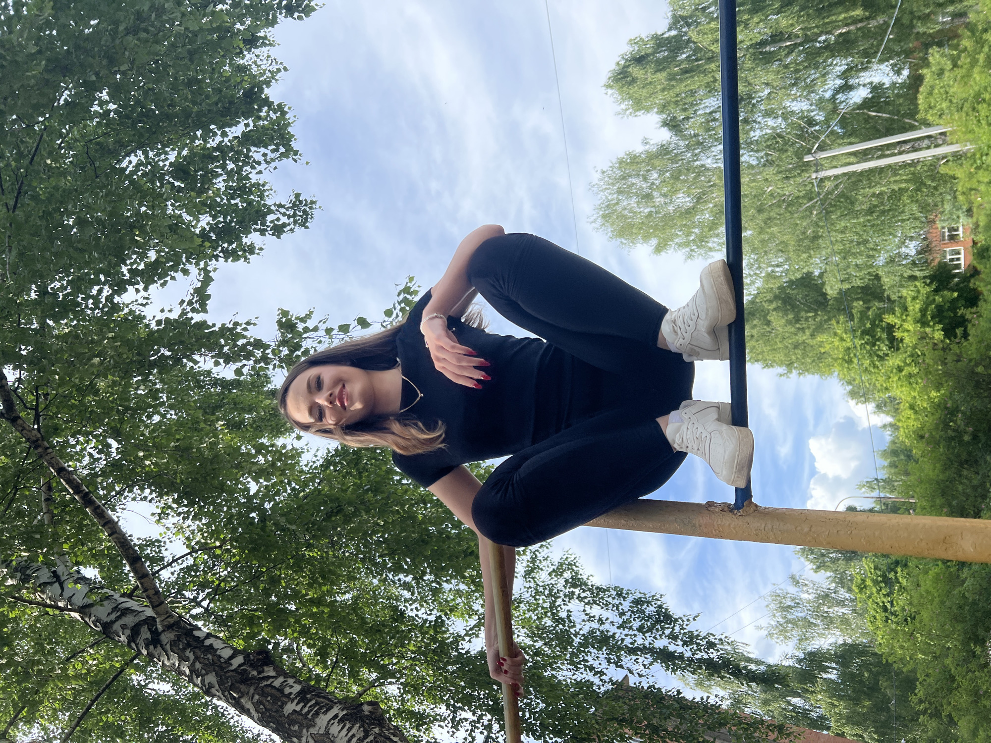 The image captures a young woman posing playfully outdoors on a bright, sunny day, likely in Noviy, Russia. She is seated on a yellow-painted or wooden horizontal bar, with her right foot, adorned in a white sneaker, propped up on a blue horizontal bar. Her body is angled, and she gazes directly at the camera with a gentle, pleasant smile. She has long, dark hair and is dressed in a black short-sleeved top, black leggings, and white athletic shoes. Notable accessories include red nail polish, a gold necklace, and a silver bracelet.
The setting is an outdoor area dominated by lush green foliage, suggesting a park, playground, or natural environment. A distinct birch tree trunk with its characteristic white and black bark is visible in the foreground to the left. Numerous green deciduous trees fill the background, reaching towards a clear to partly cloudy blue sky. In the deeper background, a building with visible windows can be faintly discerned through the trees, hinting at a nearby residential or urban area. The time of day appears to be daytime, with ample natural light. There is no visible text in the image.