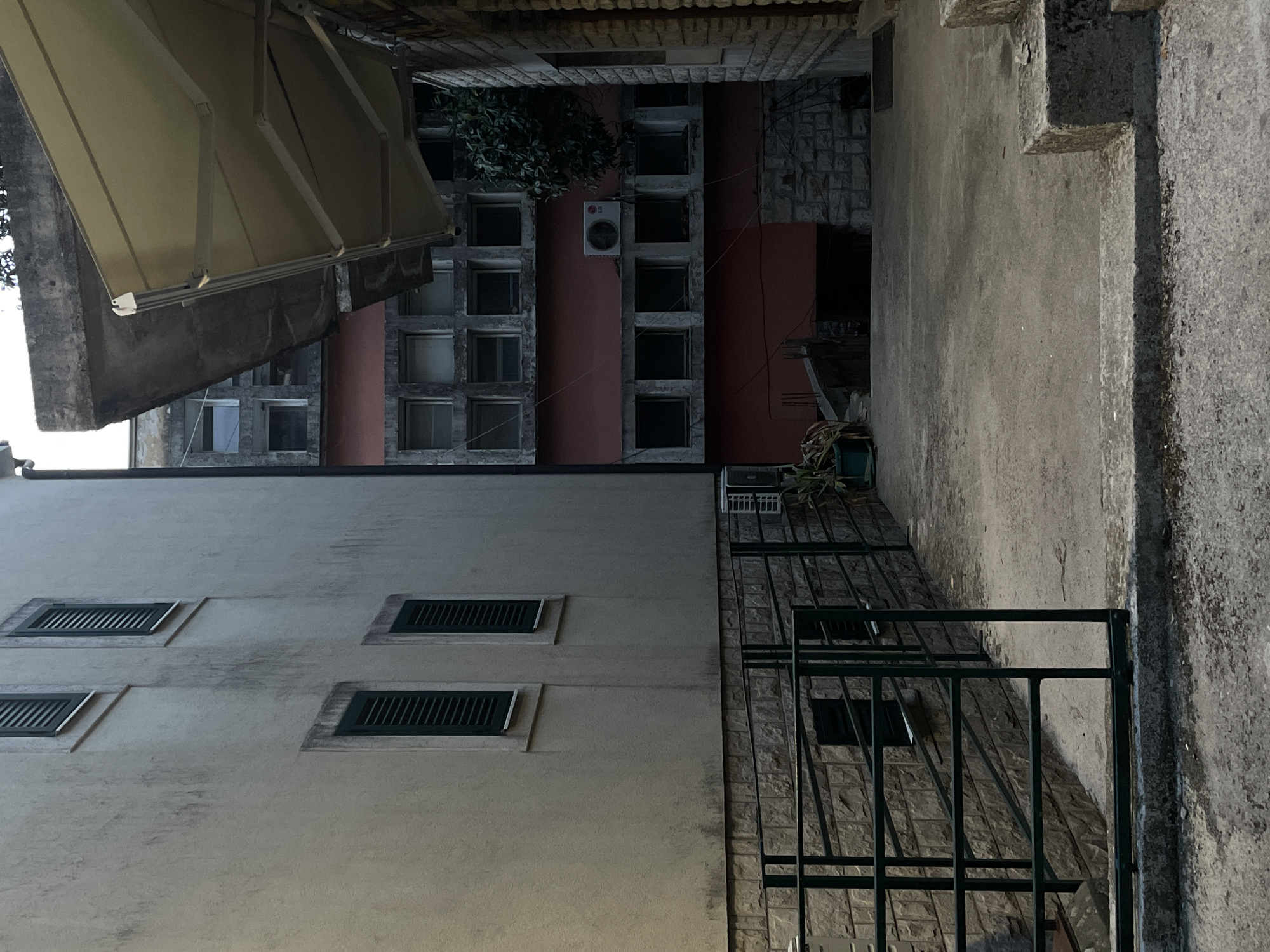 The image shows a view looking up in an outdoor area of Herceg Novi, Montenegro. A building with light-colored walls and several windows with green shutters is visible on one side. A metal railing is in the foreground. A building with a repetitive pattern of windows, some filled in, and a red-colored wall is seen in the background. An awning is present in the upper-left of the frame. The sky is not visible.