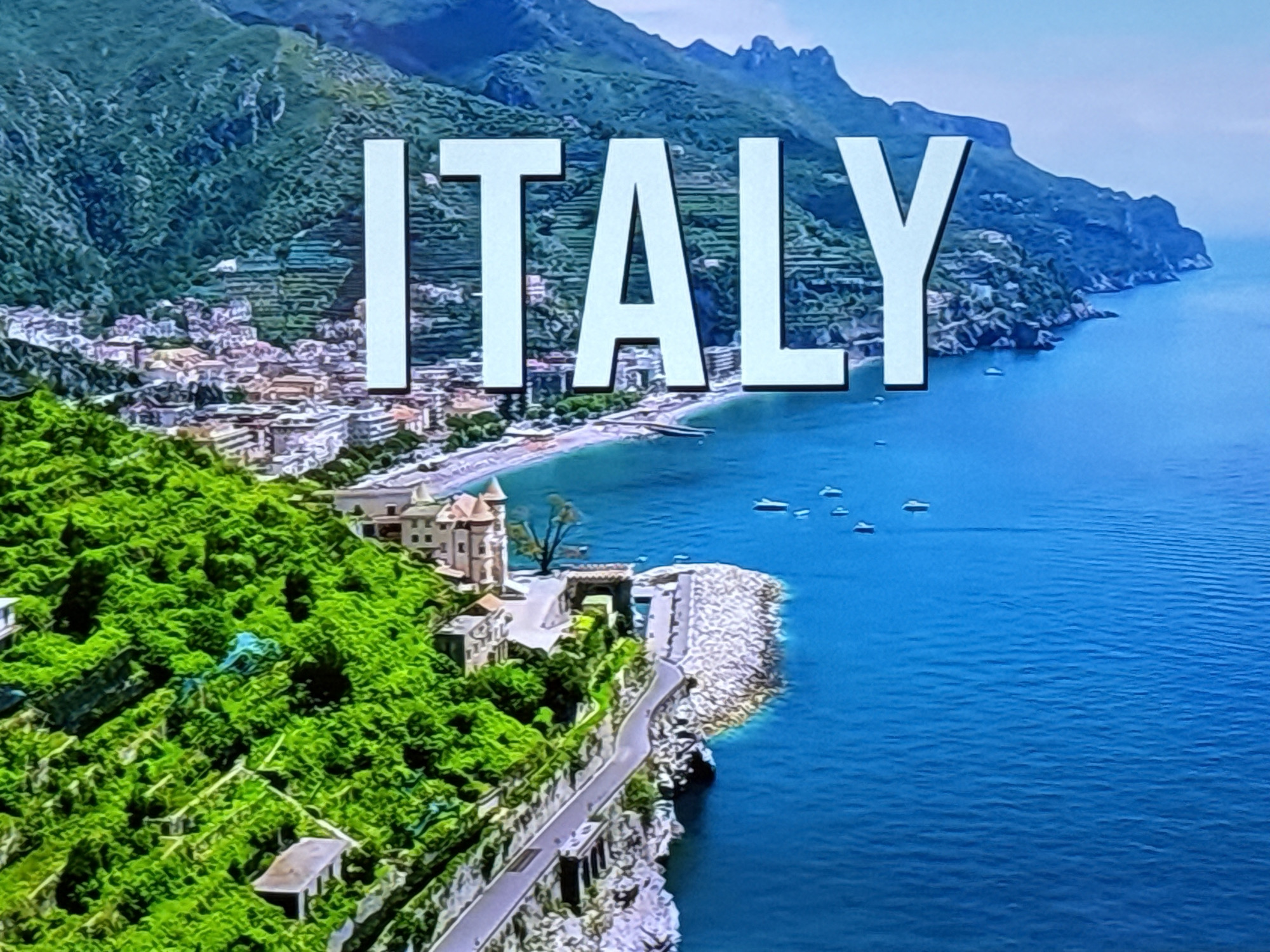 The image shows a coastal scene in Italy. The landscape includes a town with buildings nestled against green terraced hillsides, a beach, and a rocky coastline that meets the turquoise sea. Mountains are visible in the background. The word "ITALY" is superimposed on the image in large, white, sans-serif letters. Several boats are on the water.
