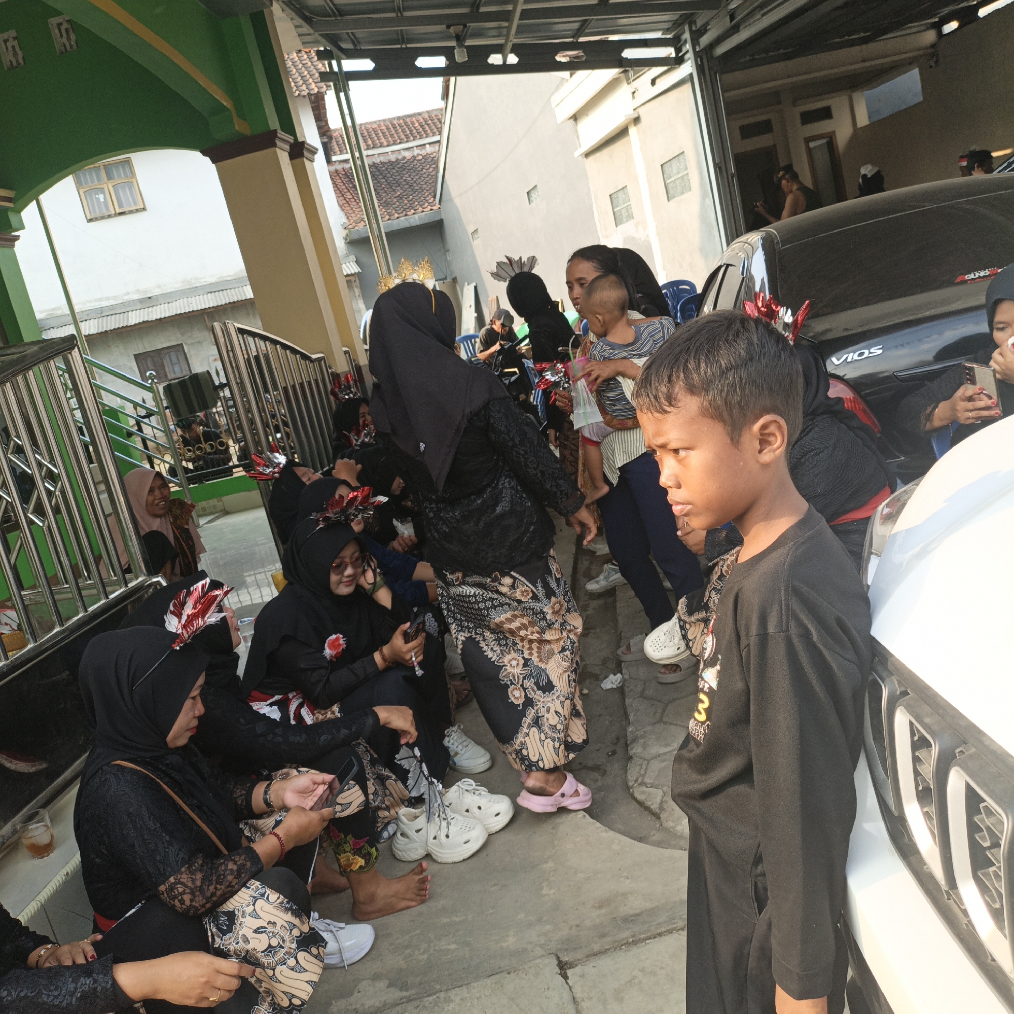 The image depicts an outdoor scene at what appears to be a residential area in Tanahbaya, Indonesia, likely a gathering or event. In the foreground, a young boy in a black long-sleeved shirt is positioned facing to the left, his profile visible. Behind him, several individuals, predominantly women wearing black hijabs and traditional patterned clothing, are seated and interacting. Some are looking at their mobile phones, while others appear to be conversing. Decorative red and white feather-like accessories are visible on some of the women's heads.
The environment includes a white car parked to the right, with the brand "VIOS" visible on its rear panel. A white vehicle is also partially visible in the foreground. The background shows residential buildings with tiled roofs and a green and yellow painted structure, possibly an entryway or part of a house. A metal gate and railing are present on the left side of the frame. The ground is a concrete surface. Sunlight suggests it is daytime. The overall atmosphere appears to be relaxed and communal.