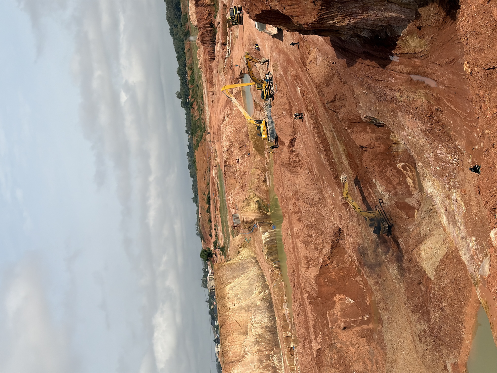 An aerial, wide-angle, eye-level shot captures a large, arid quarry excavation site under a cloudy, pale blue sky. The landscape is dominated by reddish-brown soil, with terraced layers and steep, eroded walls. Scattered throughout the quarry are several large, yellow excavators and other heavy machinery, indicating ongoing industrial activity. Puddles of murky green water have collected in depressions within the quarry. In the background, a green, tree-covered hillside rises, with some buildings and structures visible on the ridge. Several small figures, presumably workers, are dispersed across the quarry floor and on the excavated ledges, some appearing to be operating or standing near the machinery. The scene depicts a raw, industrial landscape focused on resource extraction.