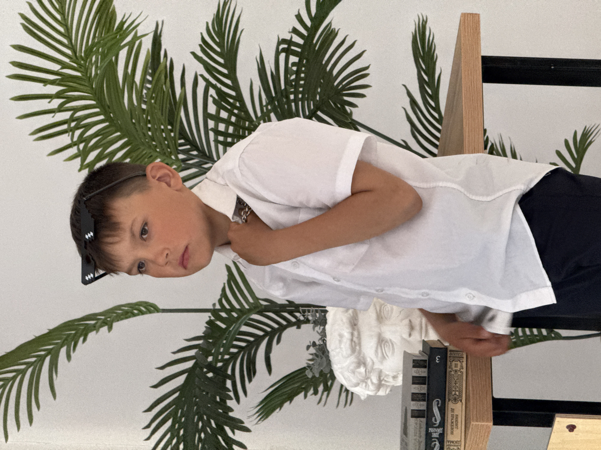 A young boy with short brown hair and sunglasses on his head is standing in front of a white wall decorated with green palm leaf patterns. He is wearing a short-sleeved white shirt and dark-colored trousers. The boy is holding his neck and appears thoughtful. Next to him, there are books stacked atop each other and a white statue. A wooden frame is also visible. The photo was taken indoors. This image does not provide enough information to verify the location in the city of Chaikovsky, Russia.