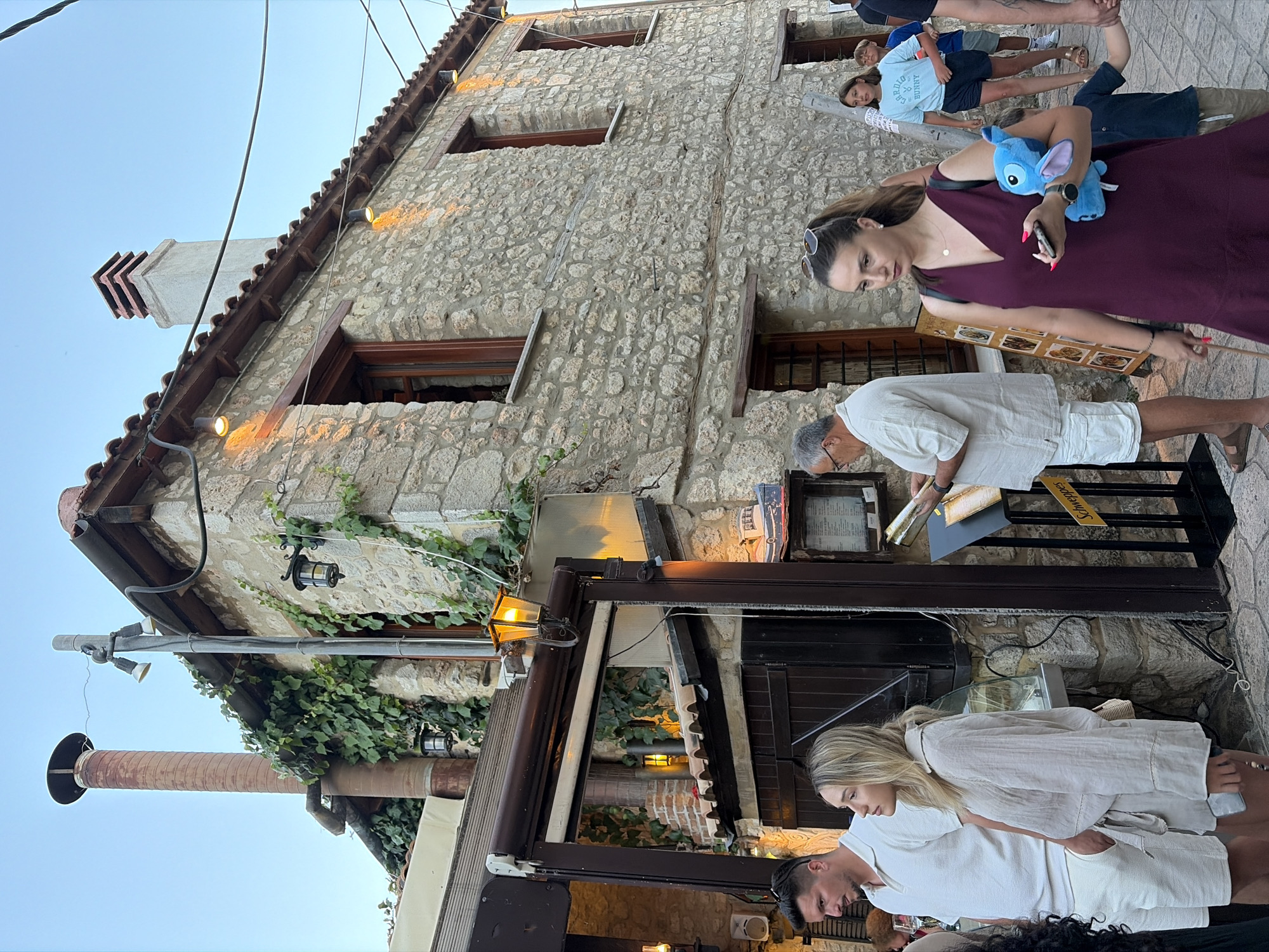 The image depicts a street scene with several people standing outside a stone building. The building appears to be a restaurant or shop, with an awning and outdoor seating. A woman in a burgundy dress is holding a phone and a plush toy, while looking down. To her right, a man in a white shirt and shorts is also looking down, possibly at a menu or table. Another woman in a white linen outfit is standing next to him, with her arm around his shoulder. In the background, a younger boy in a blue t-shirt is standing with another person. The setting appears to be a warm, possibly evening, time of day, with artificial lights illuminating the scene. The stone building and the presence of people suggest a public space in a town or village. There is some text visible on a yellow sign that reads "Schweppes". The overall atmosphere seems relaxed and casual.
