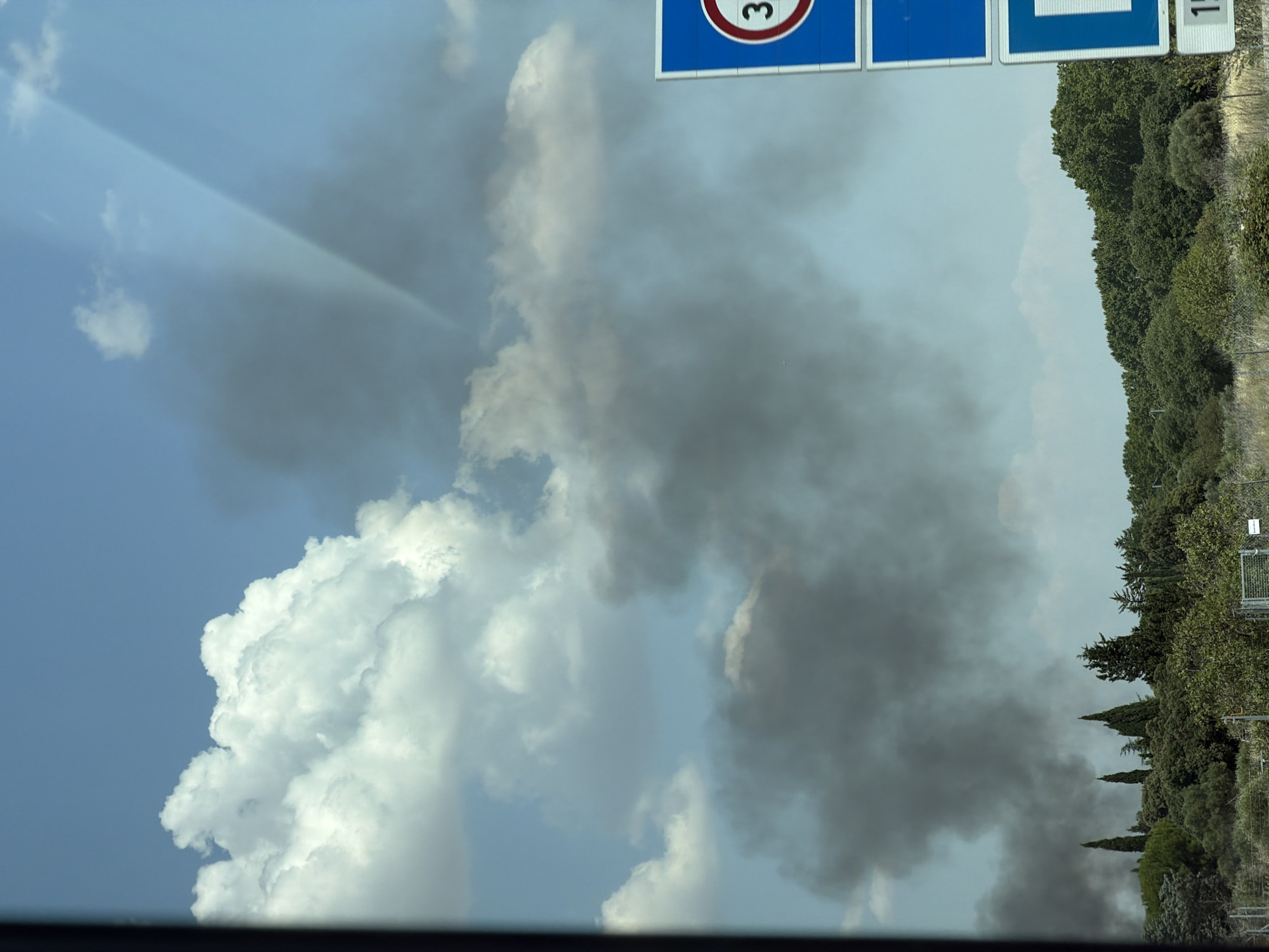 The image captures a scene in Bernis, France, dominated by a vast expanse of sky filled with both white, fluffy clouds and a thick, dark plume of smoke. The smoke appears to originate from the lower part of the image, obscuring a portion of the treeline which consists of various green trees and foliage. Two blue road signs are visible on the right side of the frame. The upper sign is rectangular with a red-bordered circle containing the number "30." Below it is another rectangular blue sign with a white shape, partially visible, and beneath that, another sign with the number "15" in black on a white background. The lighting suggests it is daytime, possibly late morning or early afternoon. There are no visible people or animals. The environment is rural or semi-rural, with dense vegetation bordering what appears to be a roadside.