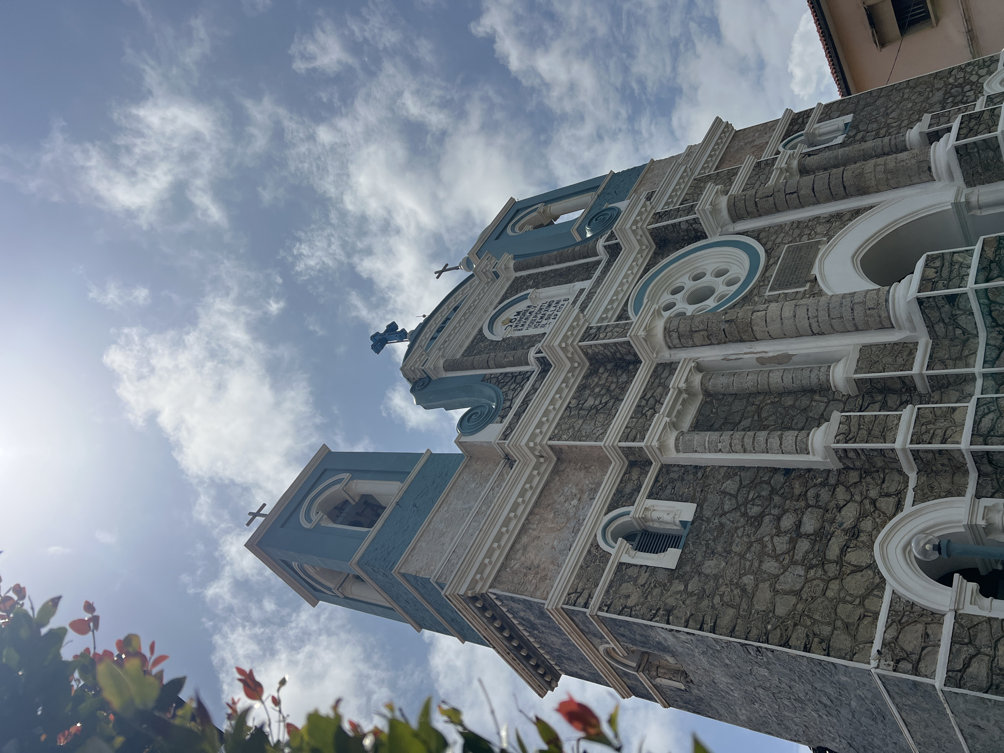 An upward shot captures the facade of a stone church in Galle, Sri Lanka, with a vibrant blue sky dotted with clouds overhead. The church's architecture is a blend of stone and white ornamental details, with two towers, one taller than the other, crowned with crosses. The taller tower features a bell opening, and the facade itself has arched windows and decorative circular elements. A plaque with text is visible on the front of the church. In the foreground, out-of-focus green leaves and red flowers add a natural element to the composition. To the right of the church, a portion of a building with an orange tiled roof is visible.