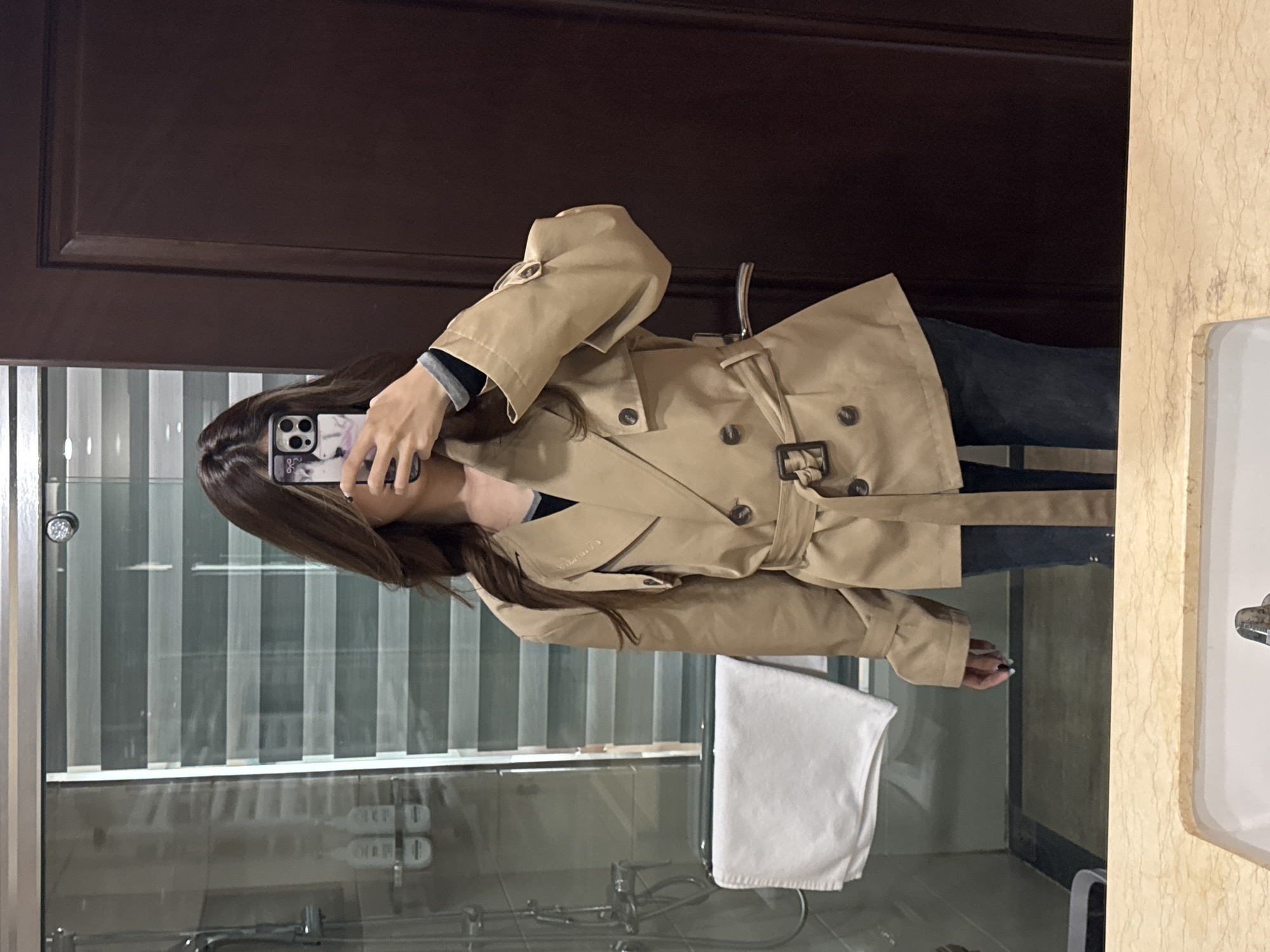 A person is holding a smartphone to take a selfie in front of a mirror in a bathroom. The person is wearing a light brown trench coat over a dark-colored top. The bathroom features a shower area with a glass enclosure. A dark wooden door is visible in the background. There is a white towel hanging near the shower. The location appears to be an indoor bathroom setting. There is no clear visual evidence that would confirm that this picture was taken in Lanzhou, China.