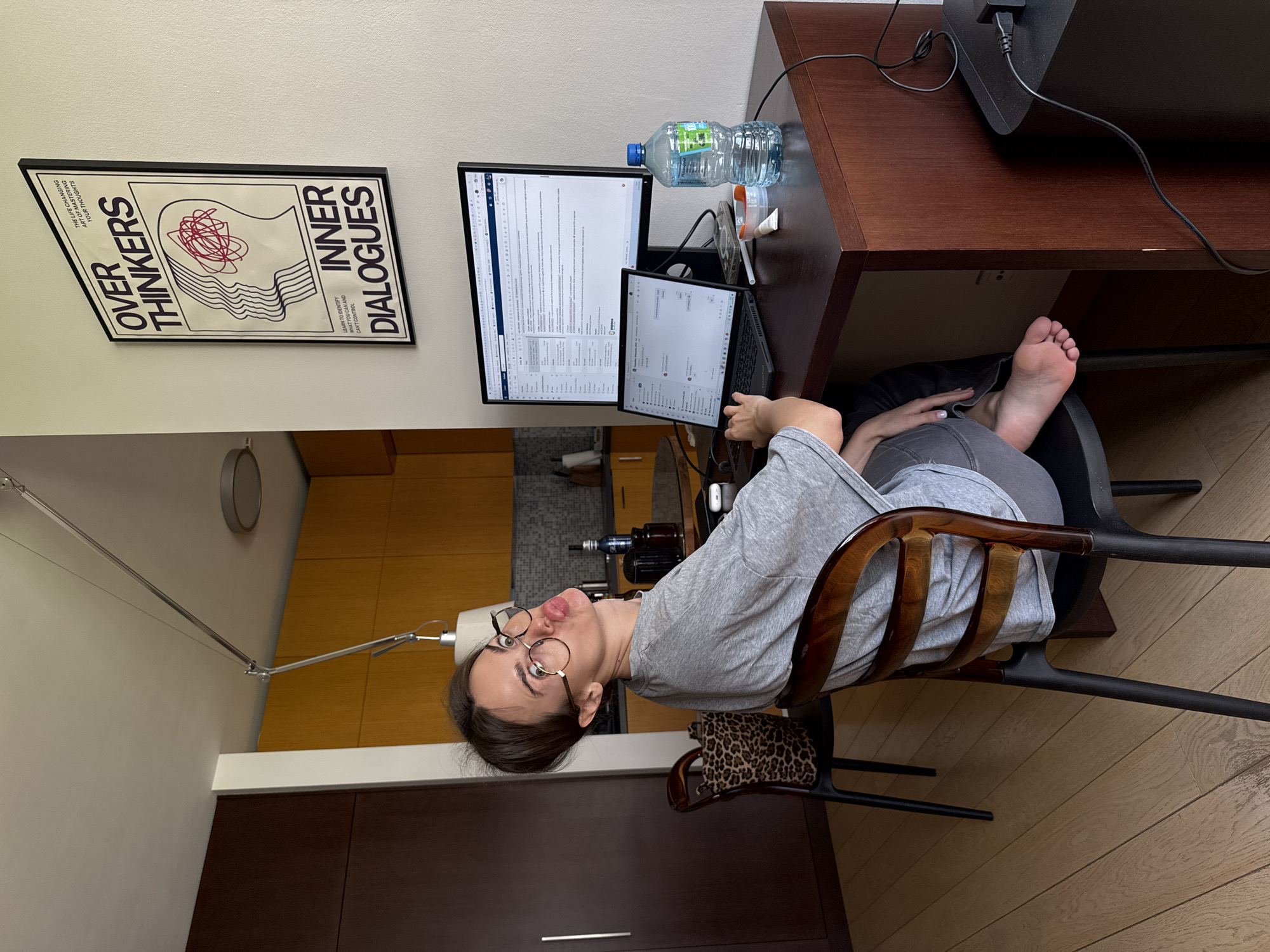 A person with glasses is seated at a desk with a laptop and monitor, with a poster that reads "Over Thinkers Inner Dialogues" on the wall. A water bottle is on the desk, and bare feet are visible under the desk. The person is in a chair, and a leopard-print bag is on the floor near the chair. The location is Unknown, Unknown.
