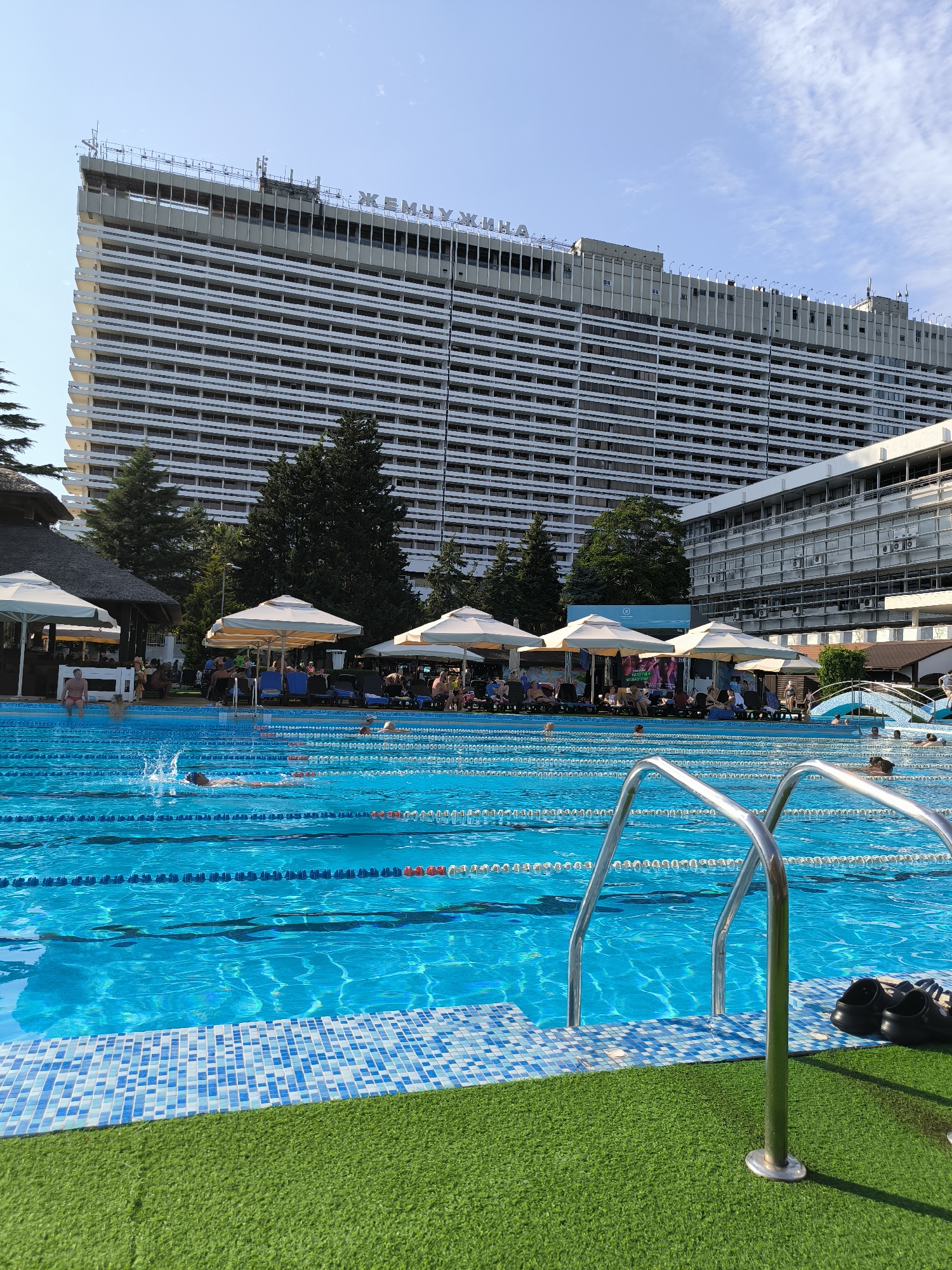 The image depicts a sunlit outdoor swimming pool area with a large hotel in the background. The hotel, identified by the Cyrillic lettering "ЖЕМЧУЖИНА" (Pearl) on its facade, is a prominent multi-story building with numerous balconies and windows. The swimming pool is a vibrant blue, divided into lanes by ropes. Several people are visible in and around the pool. Some are swimming, with one person in the foreground creating a splash. Others are relaxing on lounge chairs under umbrellas on the poolside. Adjacent to the pool is a green, artificial turf area. In the foreground, a set of chrome pool ladders is partially visible, along with a pair of black shoes placed on the turf. The sky above is a clear blue with a few wispy clouds. The environment appears to be a resort or hotel complex in Sochi, Russia, known for its coastal attractions.