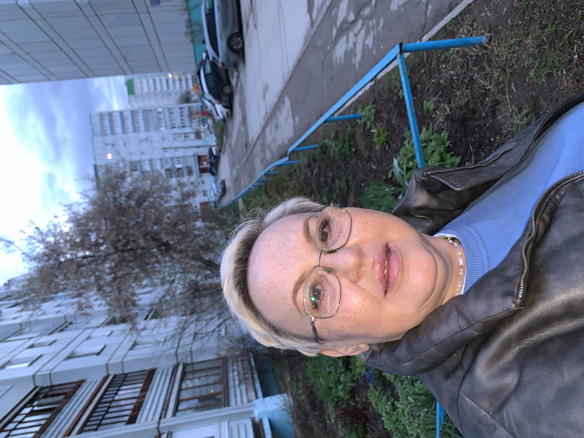A woman with short blond hair and glasses stands outside near a building in Chaikovsky, Russia. She wears a blue sweater and a black leather jacket. A blue metal railing separates her from a patch of dirt with sparse vegetation. Behind her, an apartment building rises into the sky, its windows visible. Cars are parked along a paved area. The sky is overcast.