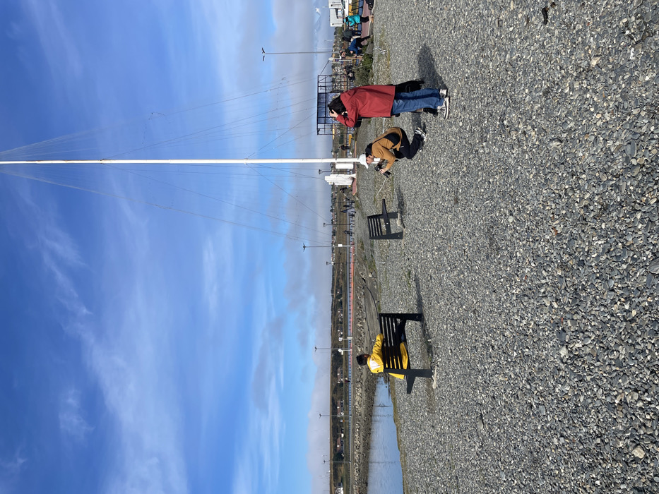 The image displays an outdoor waterfront scene in Ushuaia, Argentina, under a clear blue sky with scattered cirrus clouds. The ground in the foreground is covered extensively with grey, rounded pebbles. To the left, a calm body of water, likely a bay, is visible, bordered by a low reddish barrier or curb. A paved walkway with multiple streetlights runs along the water's edge, leading to a distant landmass with visible buildings and sparse vegetation, partially obscured by a light haze.
In the midground, a prominent, very tall, white, conical mast, secured by numerous thin rigging lines, stands upright, dominating the right-center portion of the frame. To the left of the mast, two dark-colored slatted benches are placed on the gravel. One bench, positioned closer to the water on the left, is occupied by a person wearing a bright yellow hooded jacket, seated with their back to the viewer, facing the water. The second bench, located to the right of the first, is empty.
To the right of the mast, three additional individuals are visible. One person, wearing a brown jacket, a white baseball cap, and light-colored pants, is crouched on the gravel, actively holding and aiming a black camera towards the right side of the frame. Standing immediately to their left is another person wearing a red jacket and blue jeans, with hands raised towards their head, also looking towards the right. Further in the background, near some indistinct structures on the far right, two more individuals are partially visible: one appears to be bending over in a blue garment, and another stands in a yellow garment. The scene is brightly lit by natural daylight, casting clear shadows.