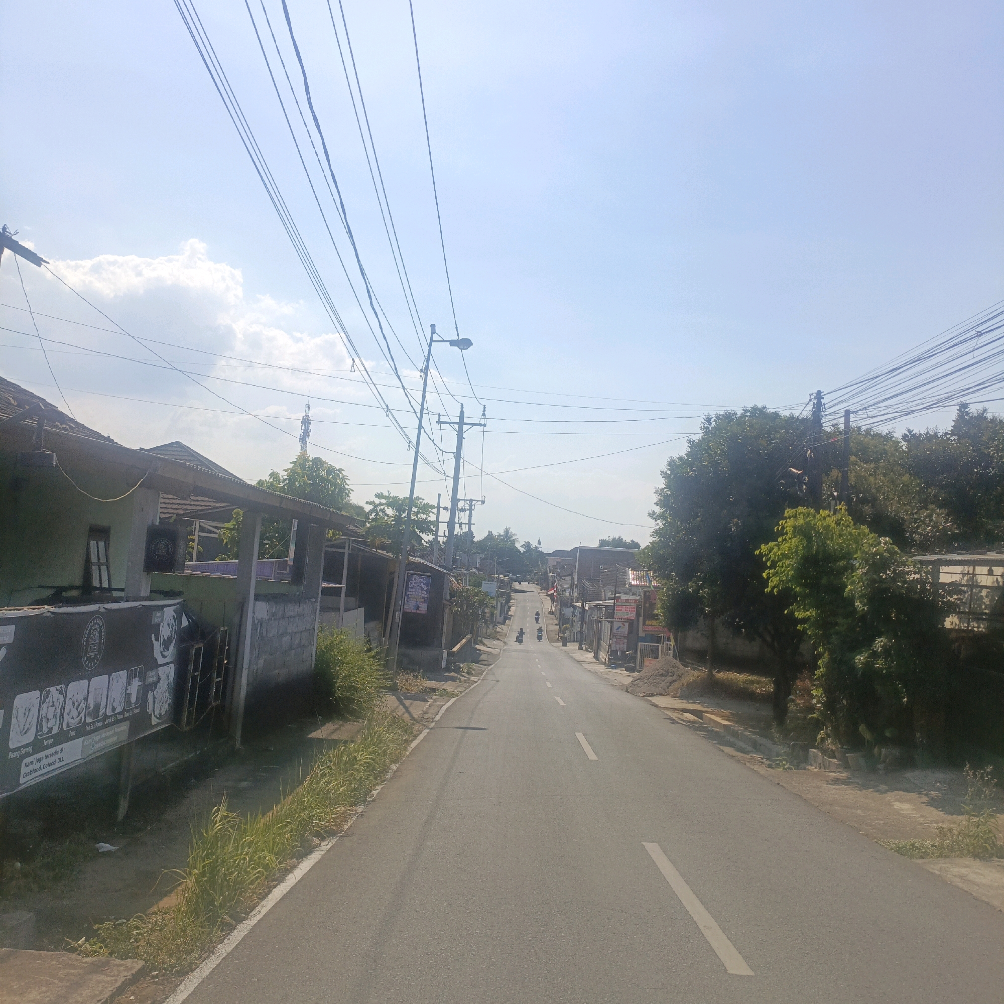 The image shows a rural road in Salatiga, Indonesia, on a clear day with a blue sky. The road is paved with asphalt and has white lane markings. On the left side of the road, there are several buildings with tiled roofs, including a green building with a window and a covered porch. A large black banner with text and images is displayed on the side of one of the buildings. Further down the road, there are more buildings, trees, and utility poles with numerous electrical wires. Several motorbikes are visible on the road, suggesting a moderate level of traffic. The right side of the road features more trees, buildings, and piles of construction materials. The overall scene depicts a typical street in a semi-urban or rural area of Indonesia.