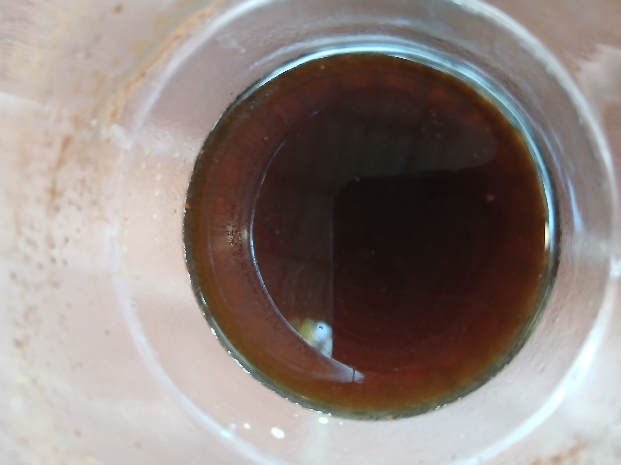The image is a close-up, overhead view of a dark brown liquid in a clear glass. The liquid has a smooth surface with some reflections. The glass appears to be cylindrical. The background is out of focus and shows a light-colored, possibly textured surface. There are small specks or bubbles visible on the surface of the liquid and on the inside of the glass. No people or specific locations within Tenggarang, Indonesia are visible.