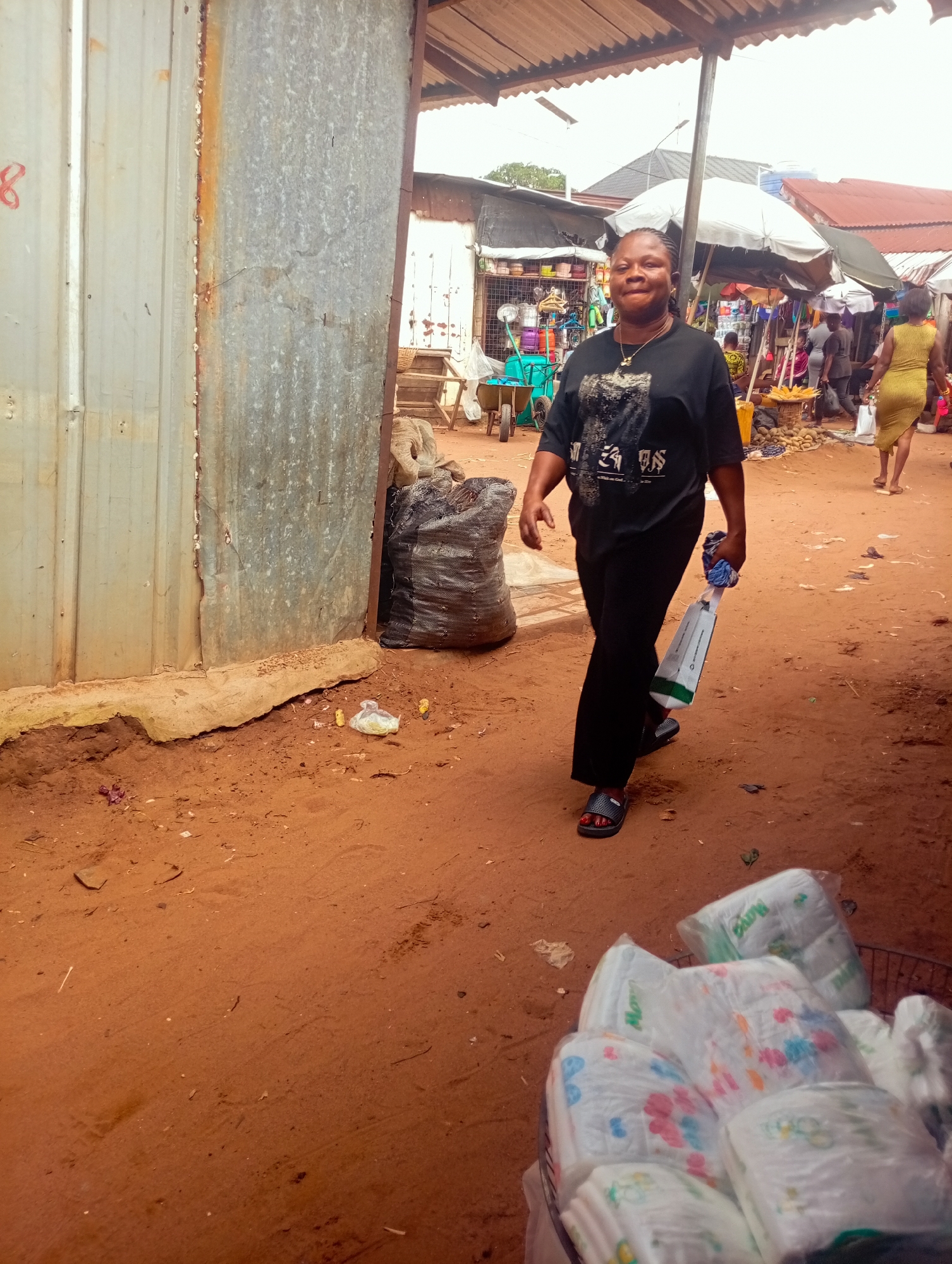 A woman walks through a bustling outdoor market in Nkpor, Nigeria. She is wearing a black t-shirt with a graphic print and black pants, accessorized with a gold necklace. She carries a white and green bag in her right hand. The ground is a reddish-brown dirt, typical of the region, with scattered debris.
In the foreground, to the right, is a woven basket filled with packages of baby diapers. Some of the diaper packages have colorful cartoon prints.
The background reveals a vibrant market scene. Stalls with awnings and umbrellas display various goods, including produce like corn and yams. Other shoppers and vendors are visible, suggesting an active marketplace. A woman in a yellow dress walks away from the camera on the right. In the distance, corrugated metal structures and roofing indicate the market's infrastructure. The sky is overcast, suggesting a cloudy day, likely in the morning or afternoon given the market activity. The overall impression is a lively, everyday market scene in Nigeria.