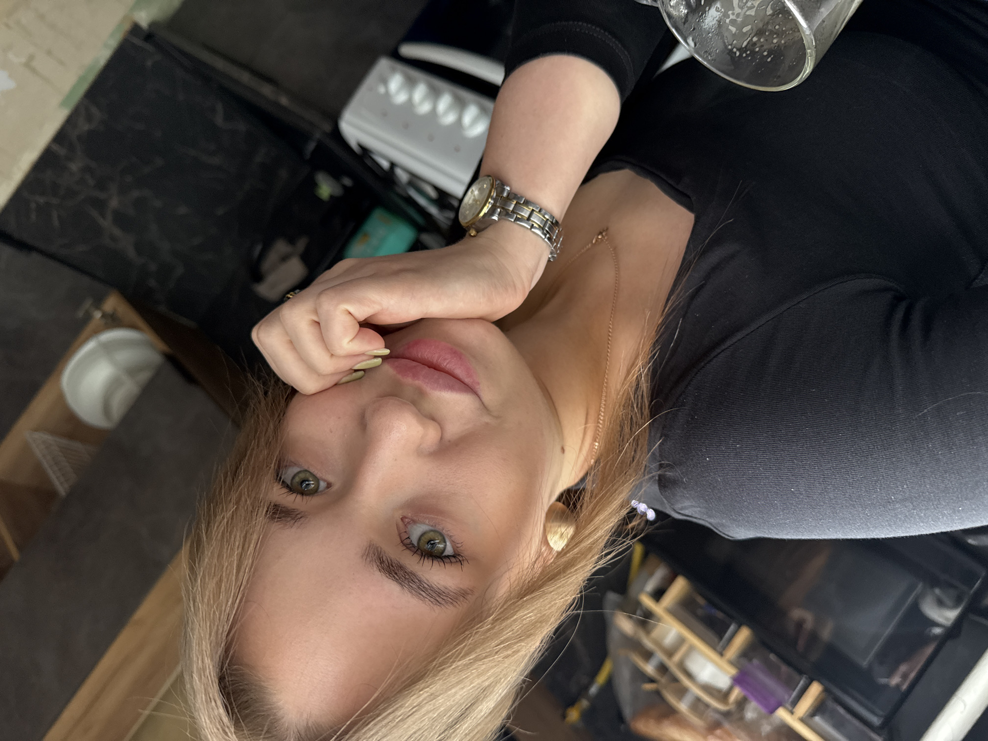 A young woman with blonde hair and green eyes is looking at the camera. She is wearing a black v-neck shirt and a gold and silver watch on her left wrist. Her right hand is resting on her chin. In the background, there are kitchen cabinets, shelves, and appliances. The image appears to be taken indoors, likely in a kitchen. The specific location in the city of Chaikovsky, Russia cannot be determined from the image alone.