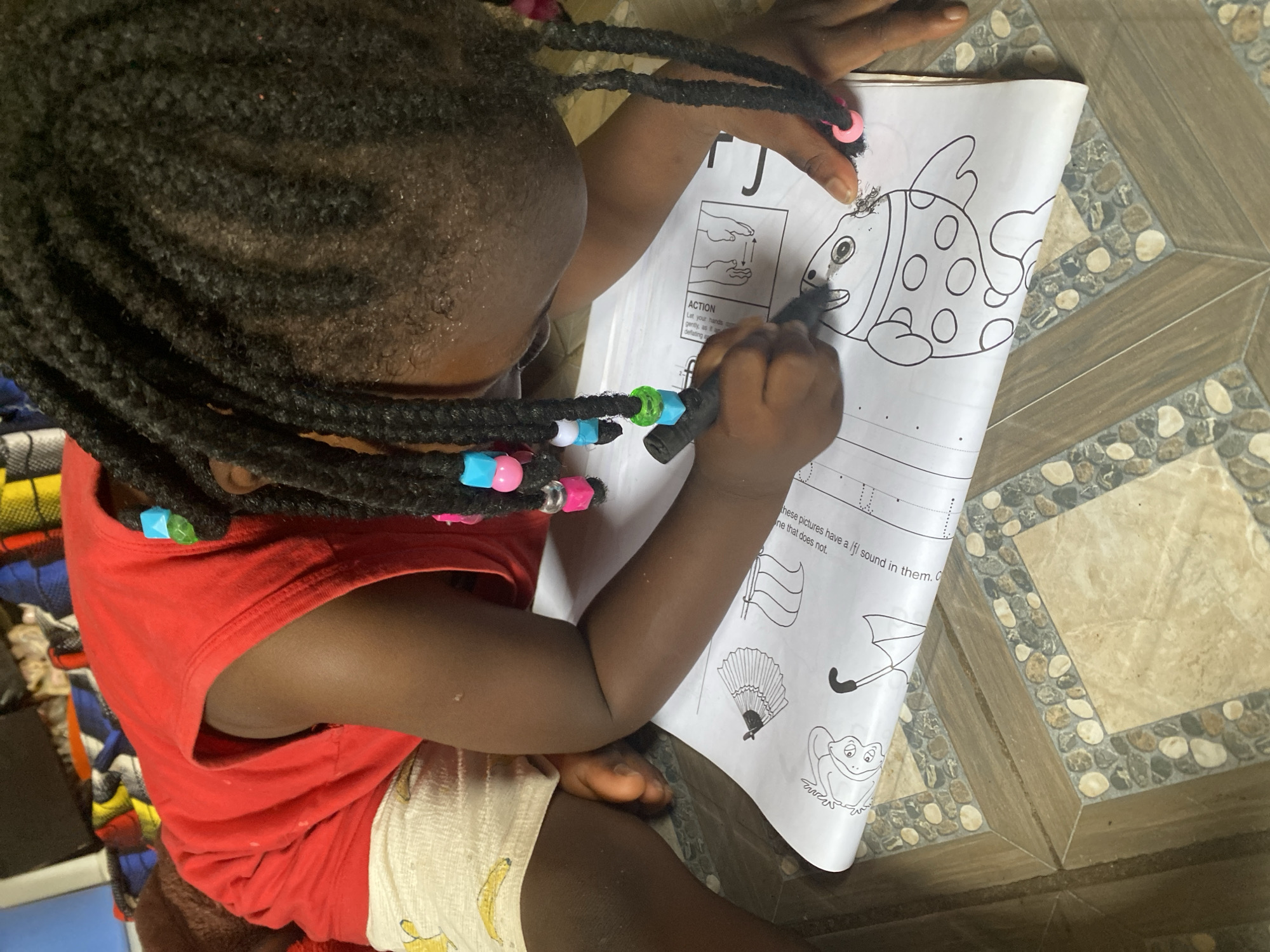 A young child with braided hair adorned with colorful beads sits indoors, likely at a home in Rido, Nigeria. The child wears a red shirt and patterned bottoms with banana designs, and is engaged in an activity involving a coloring book placed on the floor. The child is using a black marker to color a drawing of a fish. The coloring book page also contains instructions on actions and other pictures including a flag, fan, umbrella and frog. The floor is tiled with a pattern that incorporates stone-like accents.