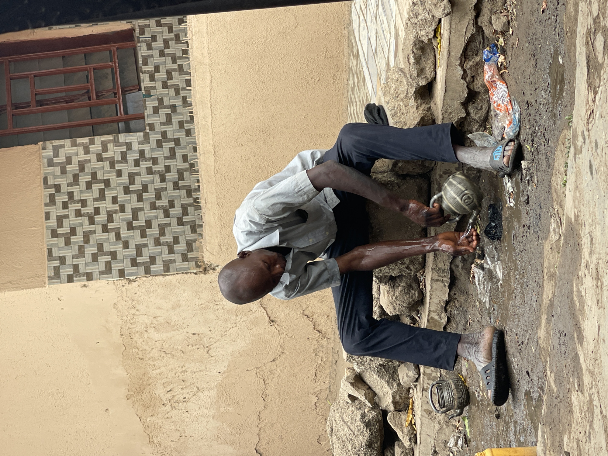 A dark-skinned man is squatting outside a building in Zaria, Nigeria, washing his hands with water from a metal pitcher. The man is wearing a light-colored shirt and dark pants. He is wearing sandals on his feet. He is squatting on a concrete surface near a wall made of stone and concrete. The ground is wet. A plastic bag lies nearby. A window with a brown frame is visible on the building behind him. The building has a light brown facade, partially covered with decorative tiles.