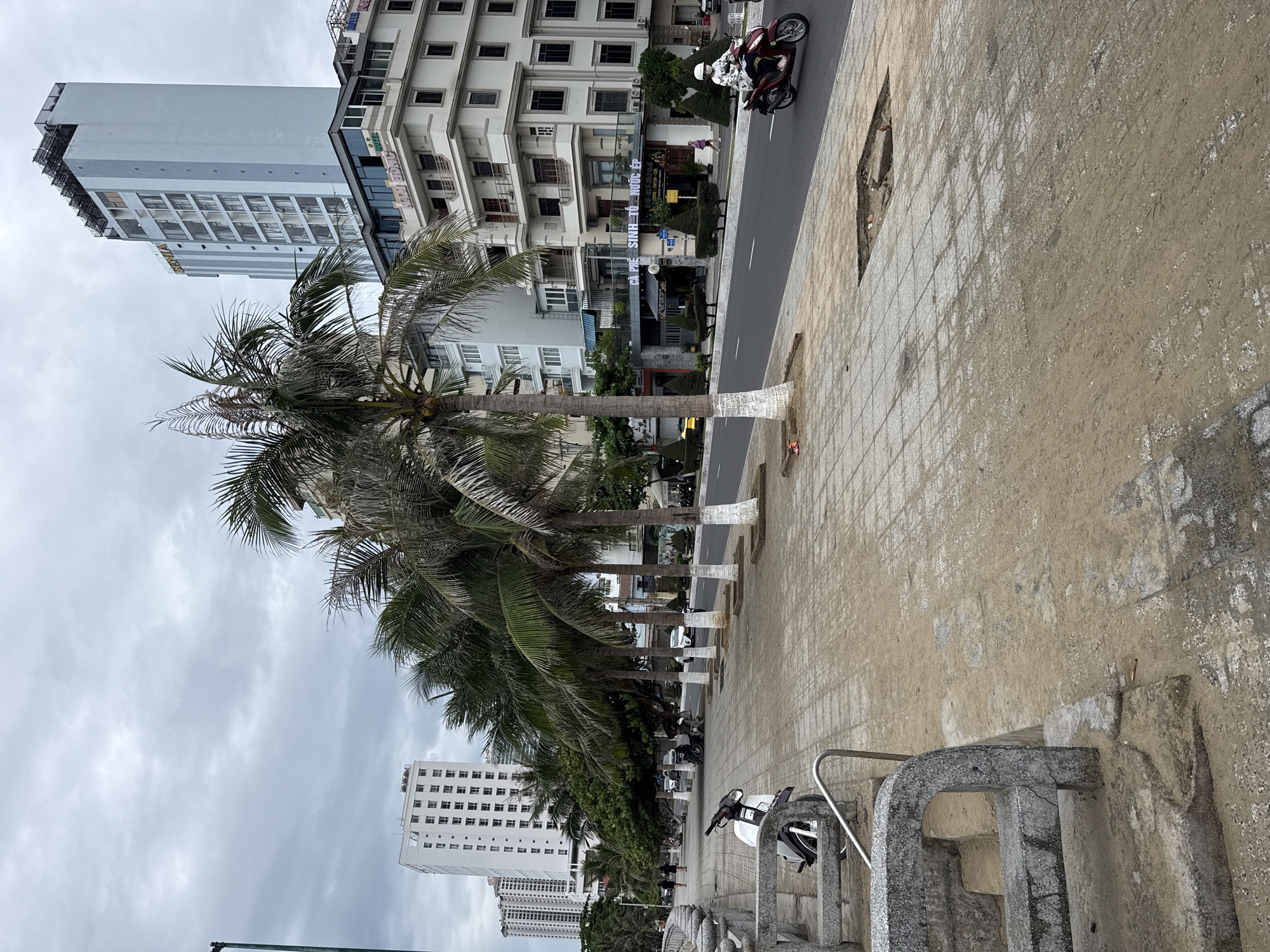 The image depicts a street scene in Nha Trang, Vietnam, likely along the coast. In the foreground, a paved sidewalk with patches of sand and a damaged section is visible, along with a stone railing and a parked white scooter. A line of palm trees with whitewashed bases runs along the sidewalk, separating it from the street. On the street, a person wearing a helmet and a light-colored jacket is riding a red scooter. In the background, there are multi-story buildings, including a tall, modern white building and other buildings with various architectural styles, suggesting a mix of hotel and commercial establishments. The sky is overcast with gray clouds. The overall impression is a busy urban environment with coastal elements.