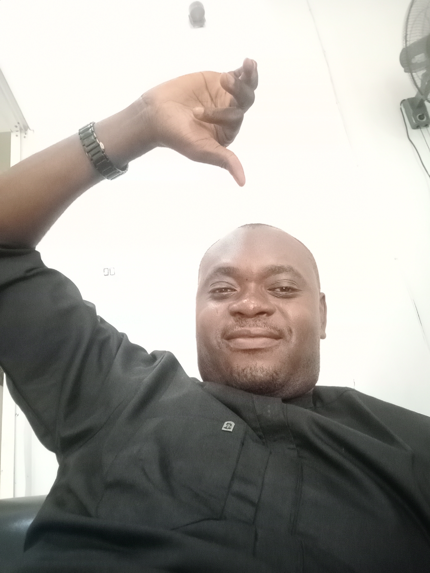 A Black adult male is depicted in a close-up selfie, looking forward with a slight smile. He has a shaved head and light stubble on his face. He is wearing a black, traditional-style tunic or kaftan, which has a small, light-colored emblem visible on the left chest area. A dark, metallic-looking watch or bracelet is worn on his left wrist. His left arm is raised, and his hand is positioned in a thumbs-down gesture above his head, directed towards the camera.
The scene is indoors, with bright white walls or ceiling dominating the background. A ceiling-mounted oscillating fan is partially visible on the upper right side of the frame. Below the man, a dark surface, likely a chair or sofa, can be seen. On the white surface behind his raised arm, a small, metallic object resembling the letters "OU" or a staple is present. The man's expression, combined with the thumbs-down gesture, suggests a casual and potentially playful or ironic mood. The photograph was taken in Gwarinpa, Nigeria.