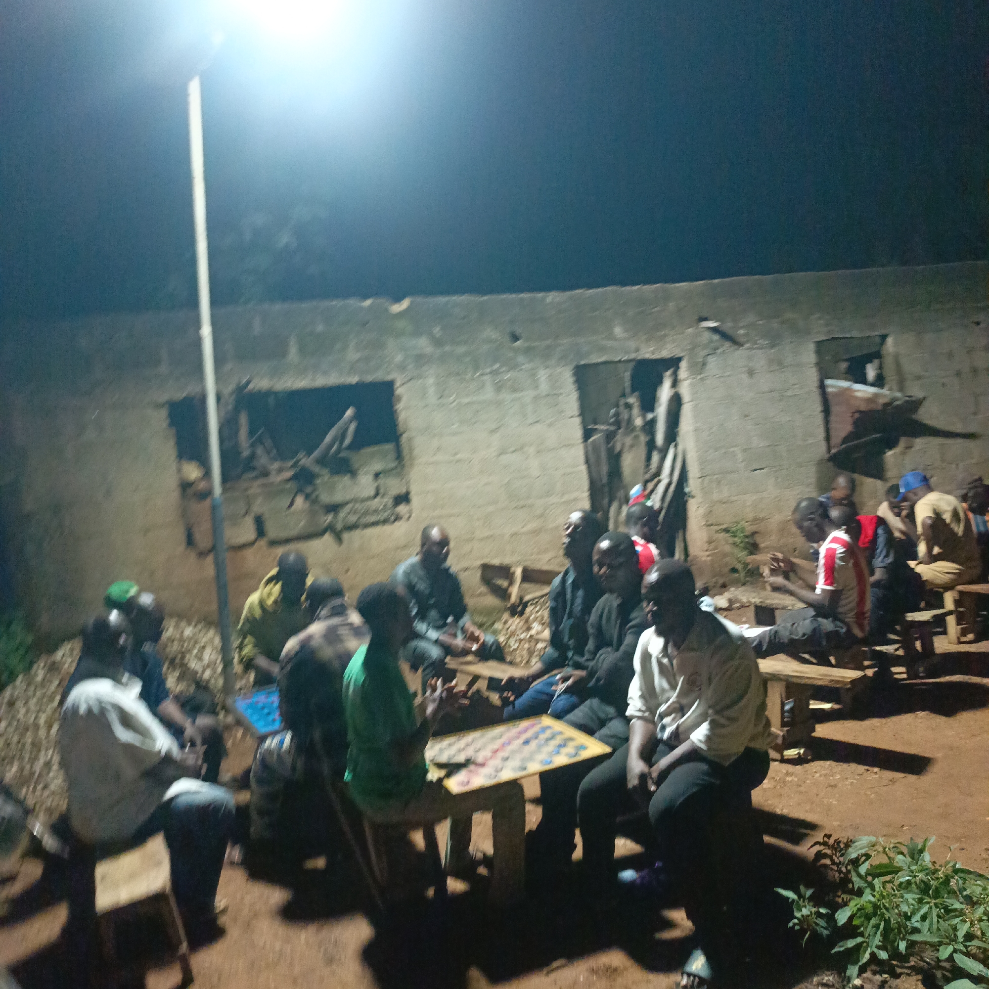In Akwanga, Nigeria, a group of men are gathered outdoors at night around a small table where they are playing a board game. The scene is illuminated by a single overhead light pole, casting long shadows on the ground. The men are seated on wooden benches and stools, their faces partially obscured by the dim light. Some are actively engaged in the game, while others observe. In the background, a dilapidated brick building with open doorways and windows suggests a rural or underdeveloped setting. The ground is covered in dirt and scattered debris, indicative of an unpaved outdoor area.