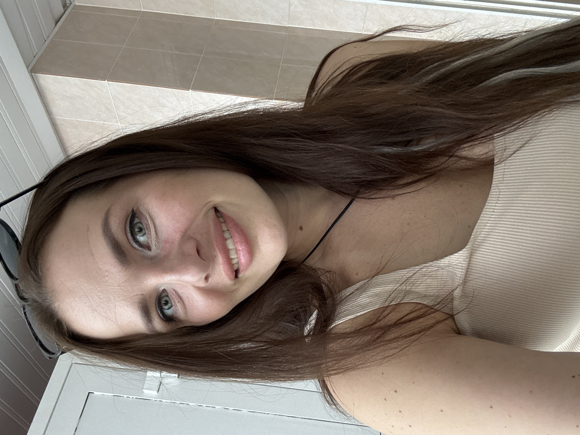A young Caucasian woman with long, dark brown hair and green eyes smiles at the camera. She is wearing a light-colored ribbed tank top. Sunglasses are perched on her head. The background appears to be an indoor setting with light-colored tiled walls. Based on the visible architectural details and general aesthetic, the location is likely an apartment or hotel room in Chaikovsky, Russia.