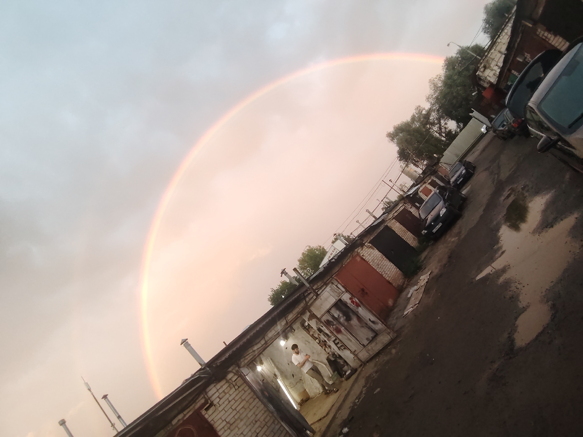 An aerial perspective, captured from a slightly tilted angle, shows a picturesque scene in Ivanteyevka, Russia, dominated by a vibrant rainbow stretching across a pale, overcast sky. Below, a row of garages creates a stark contrast with their weathered brick and corrugated metal facades. The ground is a mix of dirt and scattered puddles, reflecting the subdued light. A lone figure, dressed in a light-colored shirt and trousers, stands near the entrance of one of the garages, their posture suggesting a moment of contemplation. Several vehicles are parked in front of the garages, their shapes partially obscured by the low-light conditions and the overall tilt of the image. The scene evokes a sense of quietude, a pause in the day, bathed in the ethereal glow of the rainbow.