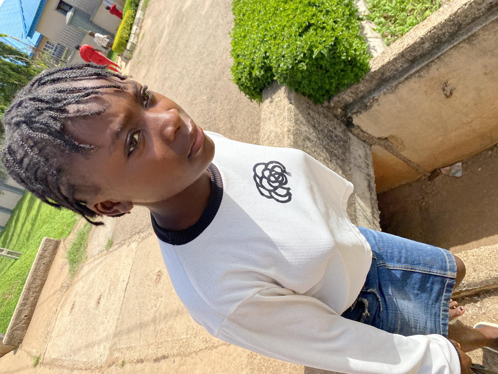 In Bukuru, Nigeria, a young girl with braided hair stands on a concrete pathway. She is wearing a white long-sleeved shirt with a black trim and a black floral design on the chest, and a blue denim skirt. Her feet are visible at the bottom of the frame. To her right, there is a dense green bush. In the background, several other individuals are seen walking, some wearing red outfits. Buildings with blue and grey roofs are visible behind them, along with green grass. The scene appears to be outdoors, possibly in a residential or institutional area.