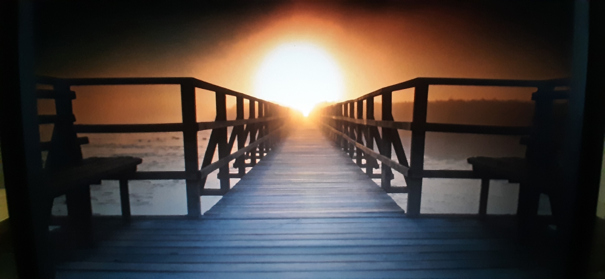 The image depicts a wooden pier extending towards a bright, hazy sun at sunset. The pier has wooden railings and benches on either side. The water beneath the pier appears calm, reflecting the warm tones of the sunset. The overall atmosphere is serene and peaceful. The location is Yessentukskaya, Russia.