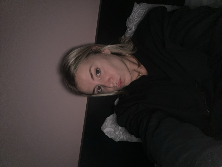 A frontal, waist-up portrait depicts a young adult female in a dimly lit indoor setting, likely a bedroom. The individual has shoulder-length, light blonde hair and light-colored eyes, looking directly into the camera with her lips puckered in a "kissing" expression. She wears a dark, long-sleeved top or jacket and a thin necklace with a small, light-colored pendant. A light-colored pillow with a faint pattern is visible behind her head and shoulder, resting against a dark headboard or wall panel. The wall above this panel is a solid light pink. The specific location within Chambœuf, France is not discernible from the visual evidence.