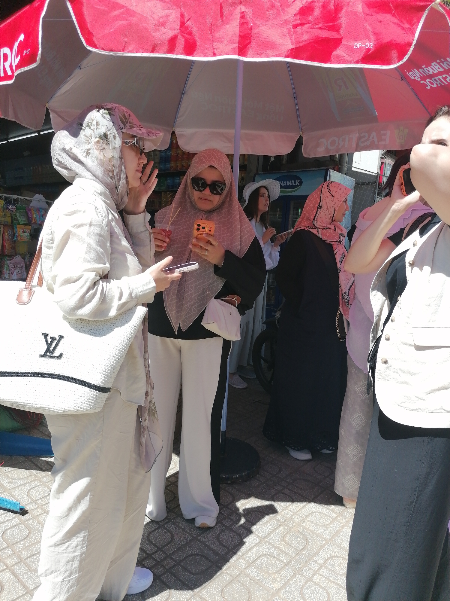 The image captures a street scene under a red and white umbrella, likely in Xuân Thành, Vietnam. In the foreground, a person dressed in light-colored clothing, including a headscarf, holds a white tote bag with a "LV" logo. To their right, another person in a light brown hijab and sunglasses is holding a smartphone and a drink. Behind them, a third person in a black abaya and a pink headscarf with a repeating pattern stands next to someone in a white hat and light-colored dress. Further back, a person in a light-colored vest is partially visible, holding a smartphone to their ear. The ground is paved with patterned tiles, and shadows are cast from the umbrella. In the background, there are stalls with merchandise and a cooler. The overall atmosphere appears to be a warm, sunny day.