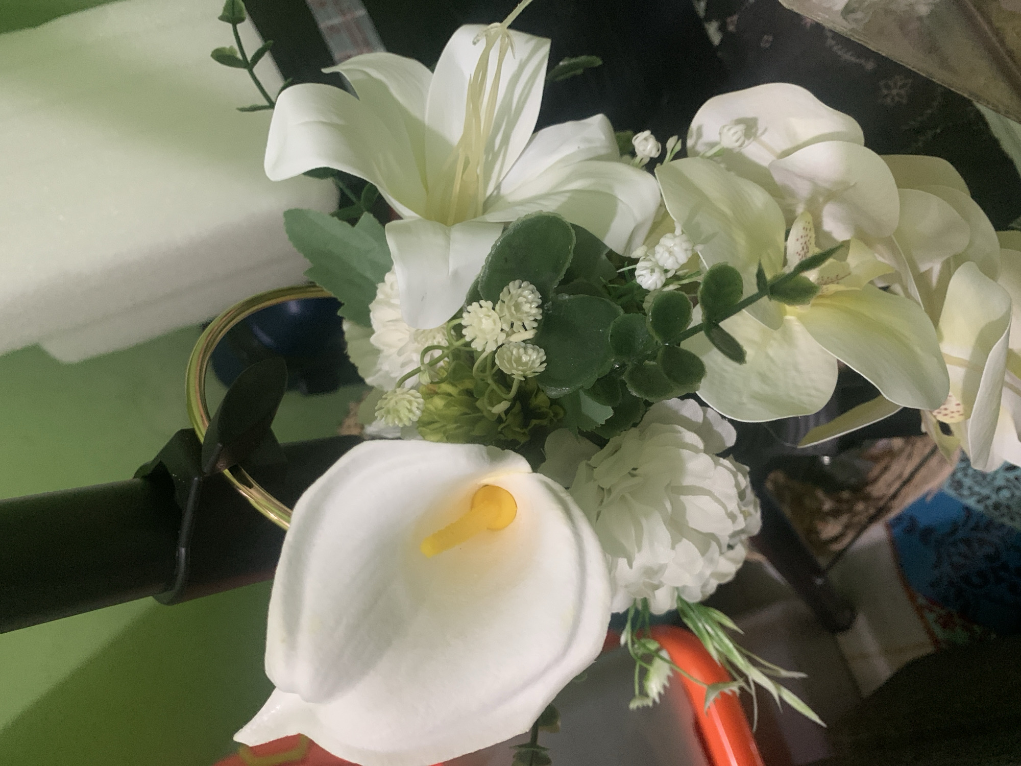 This is a close-up, overhead shot of a bouquet of artificial white flowers, primarily calla lilies and orchids, accented with greenery and small white filler flowers. A large white calla lily with a prominent yellow stamen is in the foreground, positioned slightly to the left. Behind it, several white orchids with subtle yellow markings are visible, along with a large white lily that has its stamens extending upwards. The flowers are arranged in a cluster, with various shades of green leaves and delicate sprigs interspersed.
The bouquet appears to be attached to a black metal rod with a gold ring or clasp, suggesting it might be part of a larger decorative arrangement or a prop. The background is dark and out of focus, with hints of textured fabric in blues and browns, and a bright green surface on the left, possibly a wall or table. The lighting is dim, creating shadows and highlighting the textures of the petals. There is no discernible activity or event taking place, and no visible text. The overall impression is of a still life composition focusing on the beauty of the artificial floral arrangement. The location context of Kuala Pembuang, Indonesia, is not visually evident in the image itself.