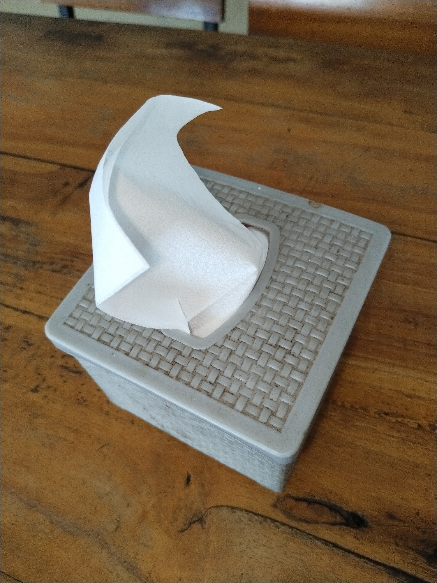 The scene is located in Matangkruet, Indonesia.
An overhead, angled photograph captures a rectangular, light gray plastic tissue box resting on a polished wooden table. The lid of the tissue box features a distinct, textured woven or basket-weave pattern. A single white tissue is partially extracted from the central oval opening on the lid, standing upright and forming a gentle, upward curve. Minor dust or debris is visible within the textured pattern of the box's lid. The wooden table displays a warm brown color with visible grain patterns and several darker knots and imperfections, suggesting a natural wood surface. The background is out of focus, showing blurred elements consistent with other wooden furniture or structures within an indoor setting. No individuals are present, and no actions or interactions are depicted.