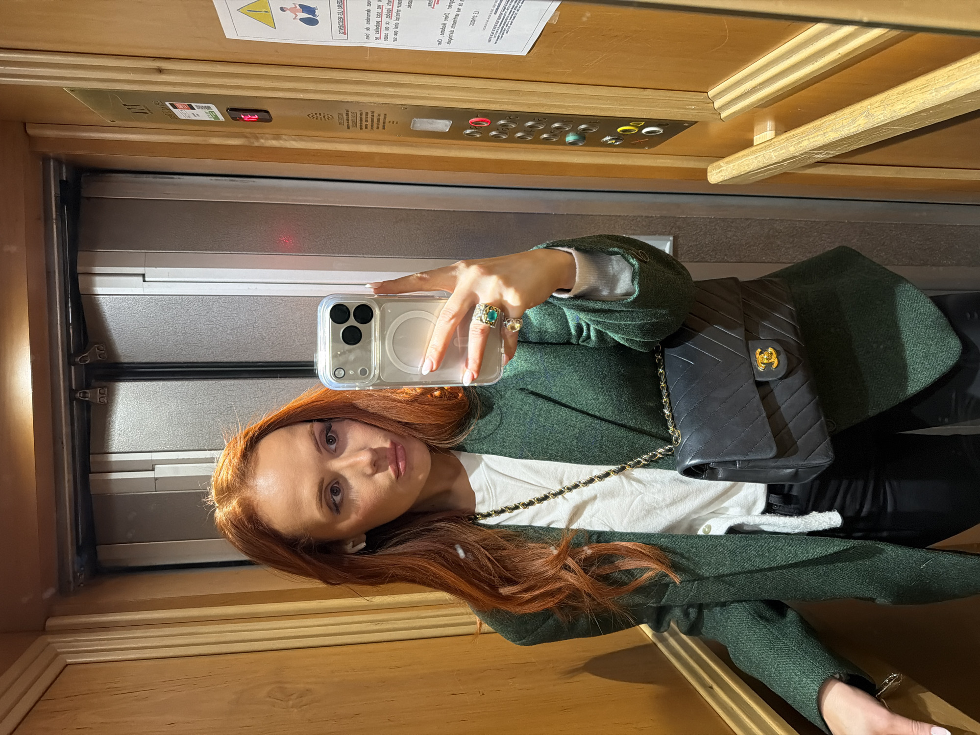 The image appears to be a selfie taken in an elevator. The woman is taking a photo of herself in the mirror. She has long, red hair and is wearing a green blazer over a white shirt. She's holding an iPhone in her hand with manicured nails and rings on her fingers. A black Chanel purse with a gold chain is slung over her shoulder. The elevator has a wood finish, with a metal door and a panel of buttons. There are signs posted above the elevator controls. I am unable to determine the location within Paris, France from the image.