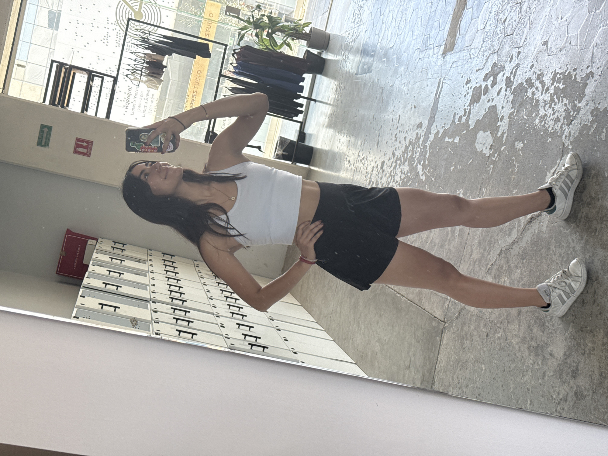 A young woman is captured in a mirror selfie, standing with one hand on her hip and looking towards her phone, which she holds up to take the photo. She is wearing a white crop top, black shorts, and white sneakers with black stripes. Her long dark hair flows over her shoulders. The setting appears to be an indoor space, possibly a gym or studio in Puebla, Mexico, given the visible elements and Spanish text.
To her left, reflected in the mirror, are rows of white lockers with black handles, and a red book or binder rests on top of some. Further left, signs for an exit (green with an arrow) and a fire extinguisher (red) are visible. To her right, clothing racks display various garments, and a potted plant sits near a window that reflects outdoor light and textures.
The floor has a textured, grey, and partially peeled concrete appearance. Visible text in the reflection includes a schedule: "HORARIO" (Schedule) listing "Lunes a Viernes" (Monday to Friday) from "7 am - 2 pm," and "Lunes a Jueves" (Monday to Thursday) for an evening session "5 pm - pm" (end time cut off). "Sábado" (Saturday) and "Domingo" (Sunday) are also listed without times. Other partial texts like "STUDIO" (repeated), "en linea" (online), and "Conta" (likely for Contacto) are discernible, suggesting a fitness or retail establishment. The overall impression is a daytime shot in an active commercial space.