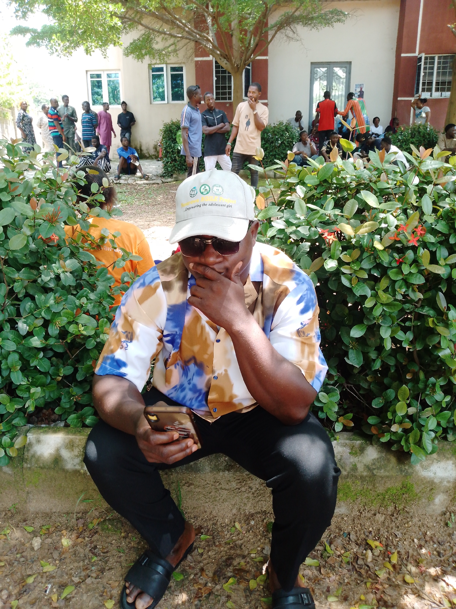 The image depicts a group of people gathered outdoors, possibly for an event or a meeting, in Akwanga, Nigeria. In the foreground, a man is seated, holding a smartphone and wearing sunglasses and a colorful, patterned short-sleeved shirt. He has his hand on his chin, suggesting contemplation or perhaps speaking on the phone. Behind him, a dense green hedge with small red flowers forms a natural barrier.
In the middle ground and background, several other individuals are visible. Some are standing and conversing, while others are seated on the ground or on a low wall. They appear to be casually dressed. The setting seems to be in front of a building with windows, and the presence of trees and foliage indicates an outdoor, possibly park-like or campus environment. The lighting suggests it is daytime, with bright sunlight casting shadows.
On the cap worn by the man in the foreground, there is visible text that reads "Empowering the Adolescent Girl." This phrase suggests that the gathering might be related to an initiative or organization focused on empowering young girls. The overall mood appears relaxed and informal. The ground is covered with soil and scattered leaves.
