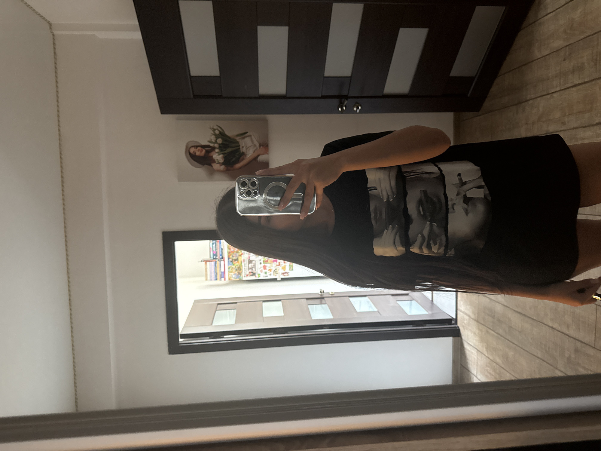 A young woman with long, dark hair stands in a room and takes a mirror selfie with her phone. She is wearing a black t-shirt with a graphic design of faces. Behind her, to the left, is an open doorway with a bookshelf visible inside. To her right, there is a dark wood door with frosted glass panels. On the wall above the door, there is a framed picture of a woman holding a bouquet of flowers. The floor is made of light-colored wood planks. The room appears to be a bedroom or dressing area. This scene is likely in Chisinau, Moldova, based on the user's prompt.
