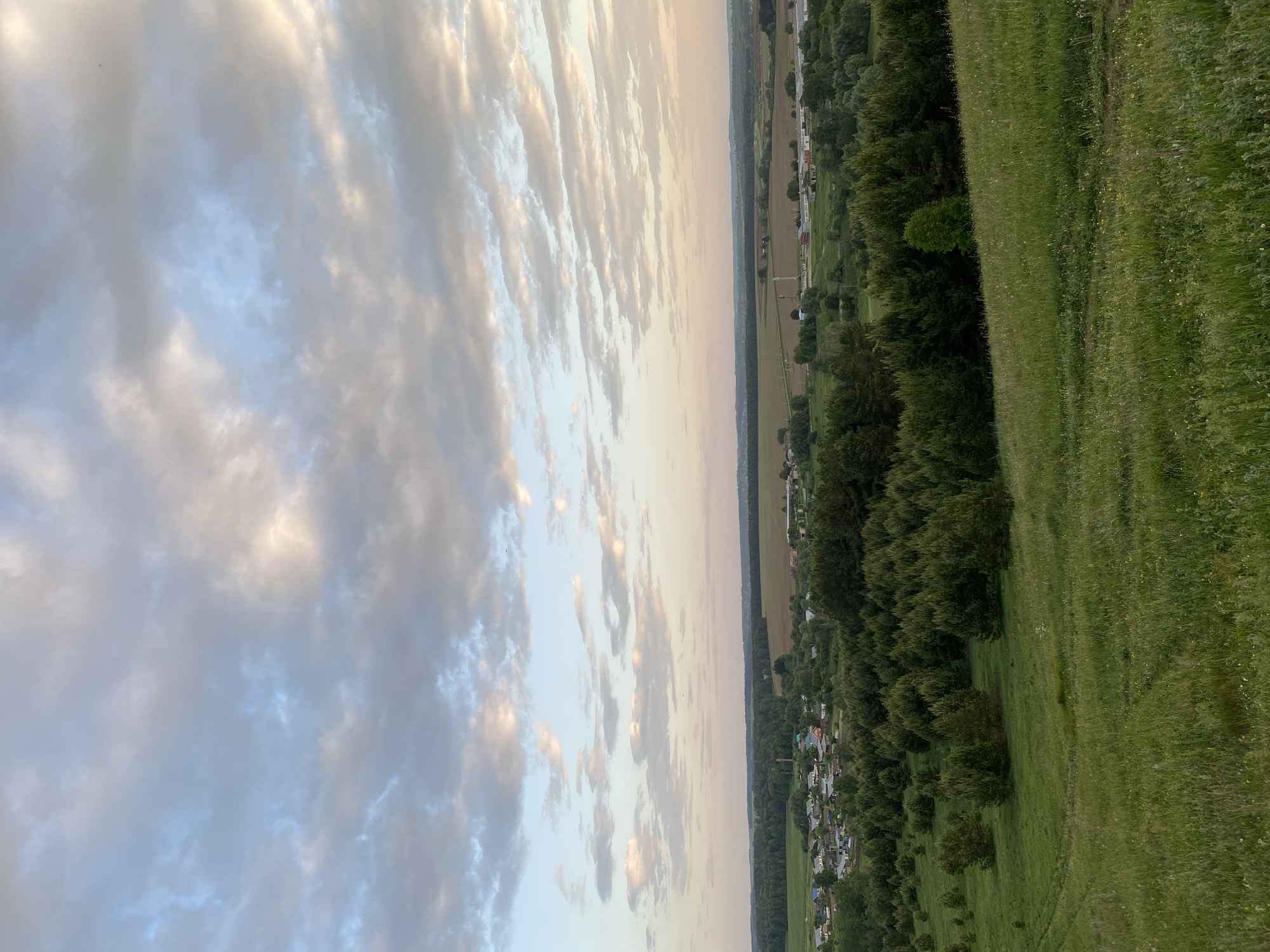 The image depicts a landscape with a clear division between sky and earth. The sky, occupying approximately half of the frame, is filled with clouds in various shades of gray and white, illuminated by what appears to be setting sunlight. The horizon line separates the sky from a lower section containing a mix of green vegetation, trees, and a small village or town. There are no people visible in the scene. The photograph gives an impression of a countryside or rural location, with the village situated along the border between a field and what seems to be the beginning of a hillside. The lighting suggests the image was taken either at dawn or dusk.