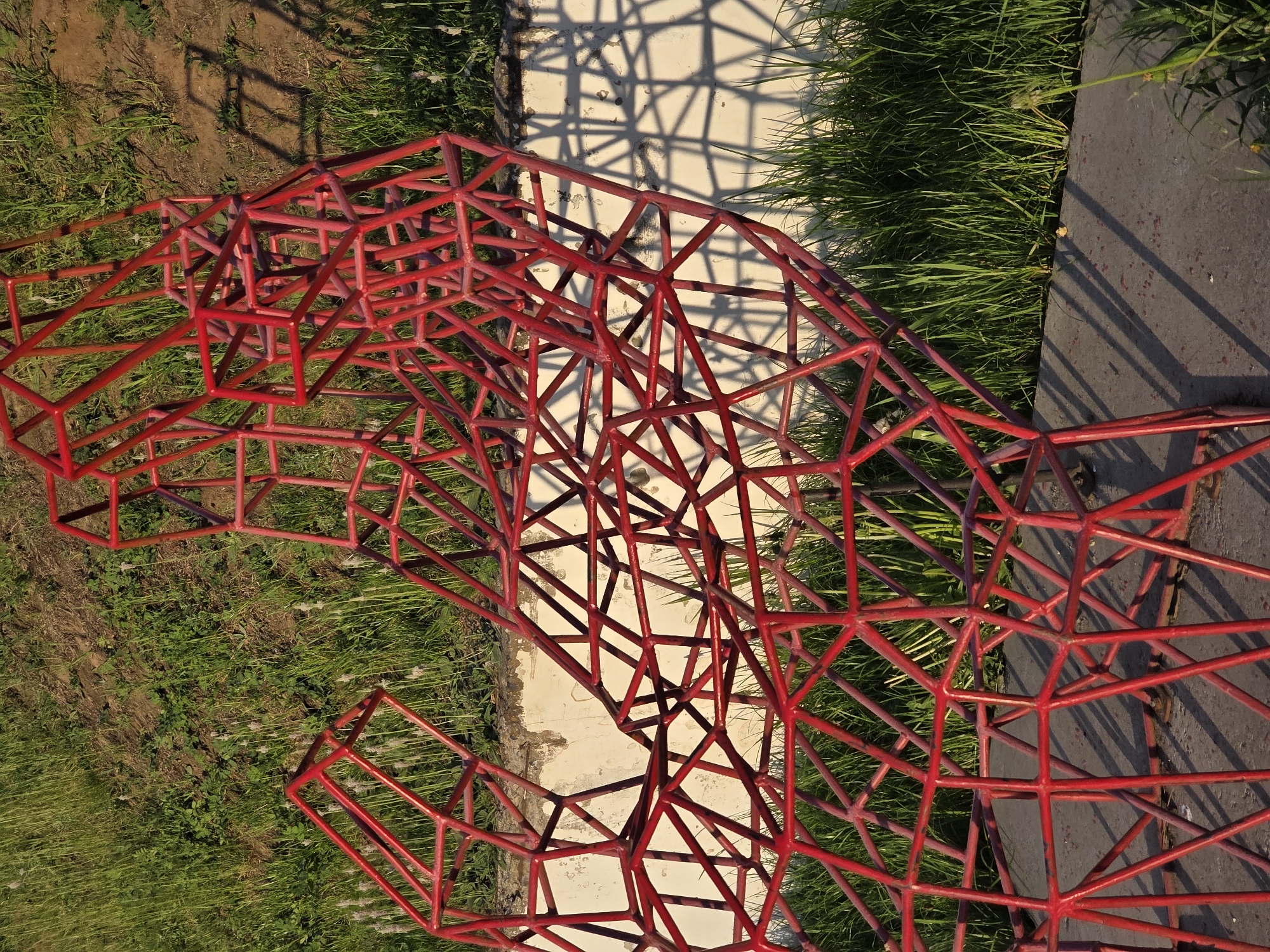 The image shows a red, three-dimensional, geometric metal structure, possibly a playground climbing frame, situated on a grassy area. The structure's shadow falls on a light-colored concrete surface adjacent to the grass. The location is likely outdoors in the city of Chaikovsky, Russia. There are no people visible in the frame. The environment includes grass and concrete, suggesting a park or recreation area.
