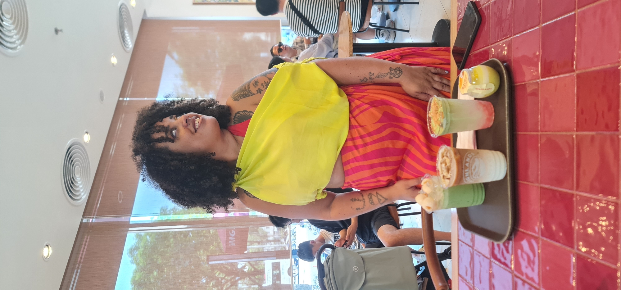 A woman with dark curly hair stands in front of a window at what appears to be a coffee shop in Đà Nẵng, Vietnam. She wears a red top, a bright yellow scarf-like garment tied at the shoulder, and an orange and pink striped skirt. Her arms are adorned with tattoos. A tray with four different types of beverages is visible on a table, one of which has a Highland Coffee logo. Other people are seated in the background. A stroller is nearby. The interior features a red tile wall and a modern ceiling with circular vents. Outside the window, trees can be seen.